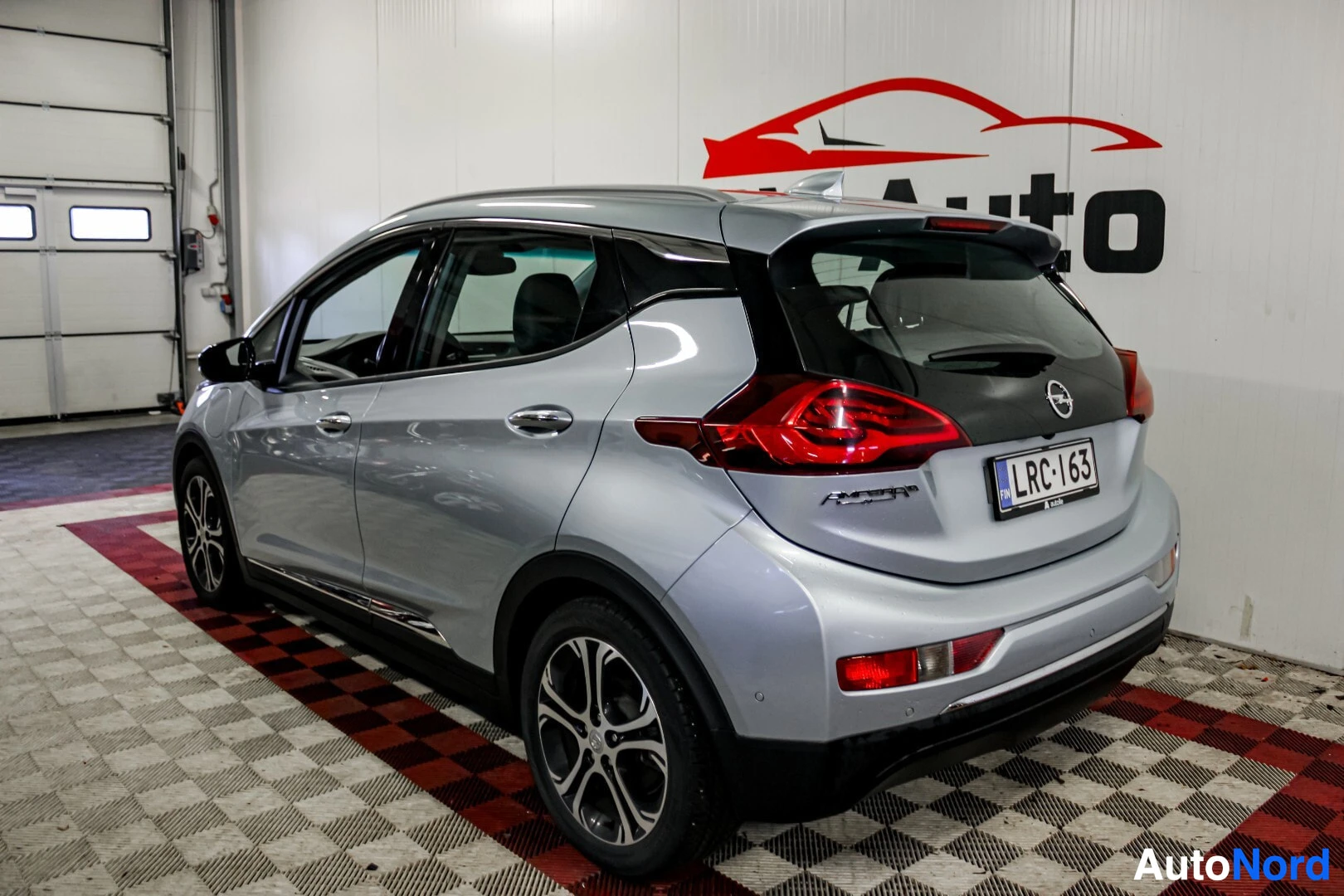 Takaviisto: Hopeanvärinen Opel Ampera-e takaapäin oikealta viistosti kuvattuna, takavalot ja takapuskuri näkyvissä. Takarekisterikilpi on näkyvillä