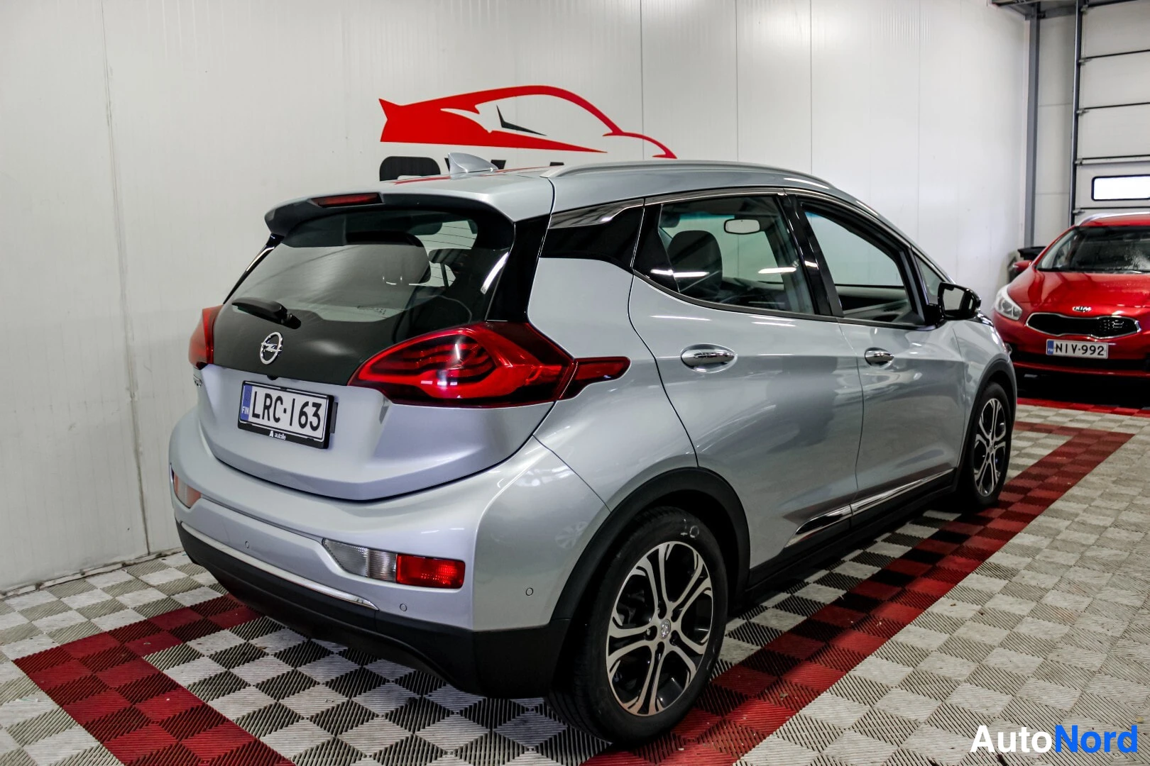 Takaviisto: Hopeanvärinen Opel Ampera-e takaapäin vasemmalta viistosti kuvattuna, takavalot ja takapuskuri näkyvissä. Takarekisterikilpi on näkyvillä