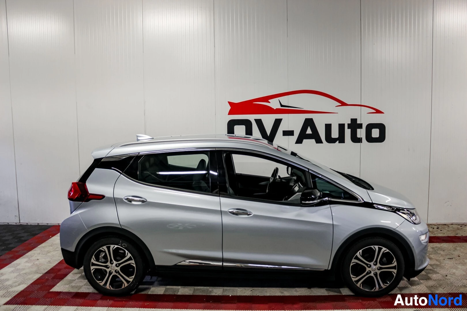 Sivulta: Hopeanvärinen Opel Ampera-e auton vasemman kyljen puolelta kuvattuna, NV-Auton logolla varustettua seinää vasten.