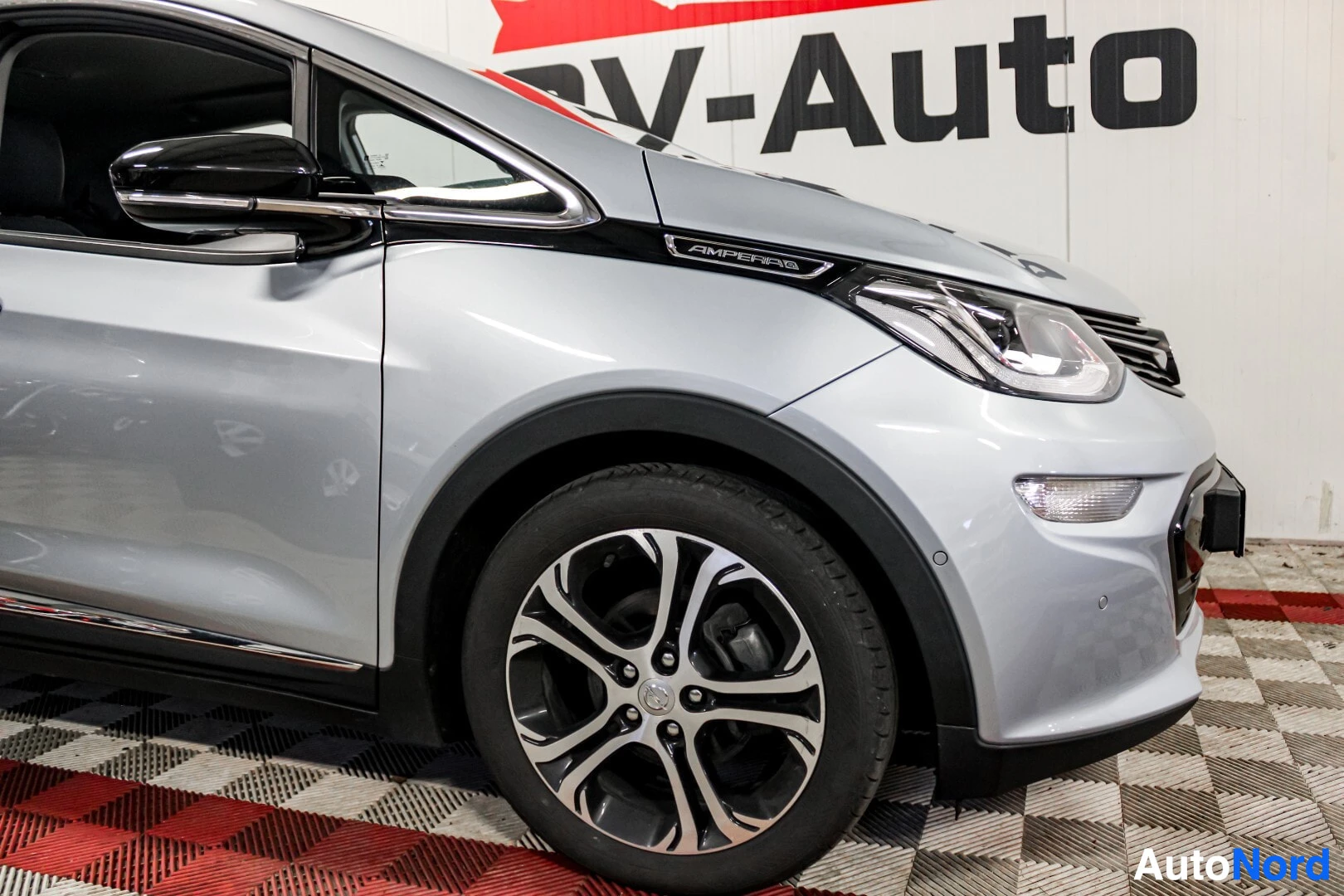 Yksityiskohta: Lähikuva auton etupäästä ja vasemmasta etupyörästä. Pyörässä on monipuolaiset kevytmetallivanteet ja Ampera-e-merkki on näkyvissä lokasuojassa.