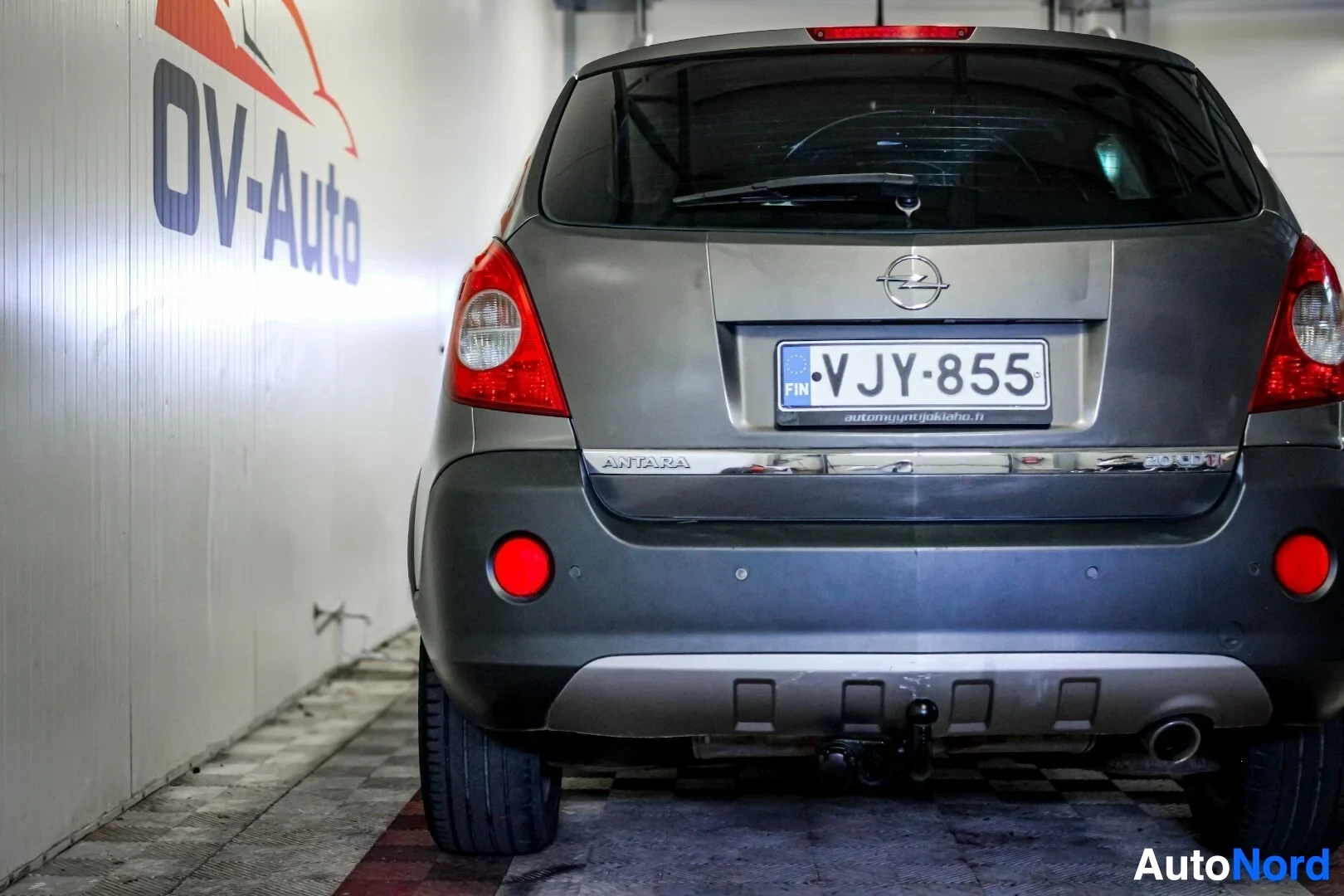 Takaa: Harmaan henkilöauton takaosa kuvattuna matalasta kulmasta. Autossa on vetokoukku ja takalasinpyyhin. Rekisterikilpi ja Opel-logo ovat näkyvissä. Oikealla puolel