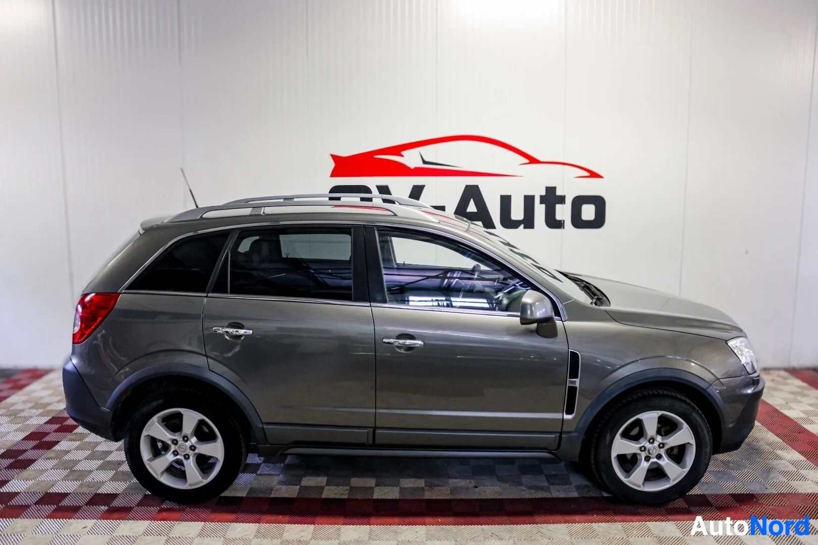 Sivulta: Harmaa henkilöauto kuvattuna vasemmalta sivulta. Autossa on kattokaiteet ja kevytmetallivanteet. Auto on pysäköity sisätiloihin, seinällä näkyy OV-Auto-logo.