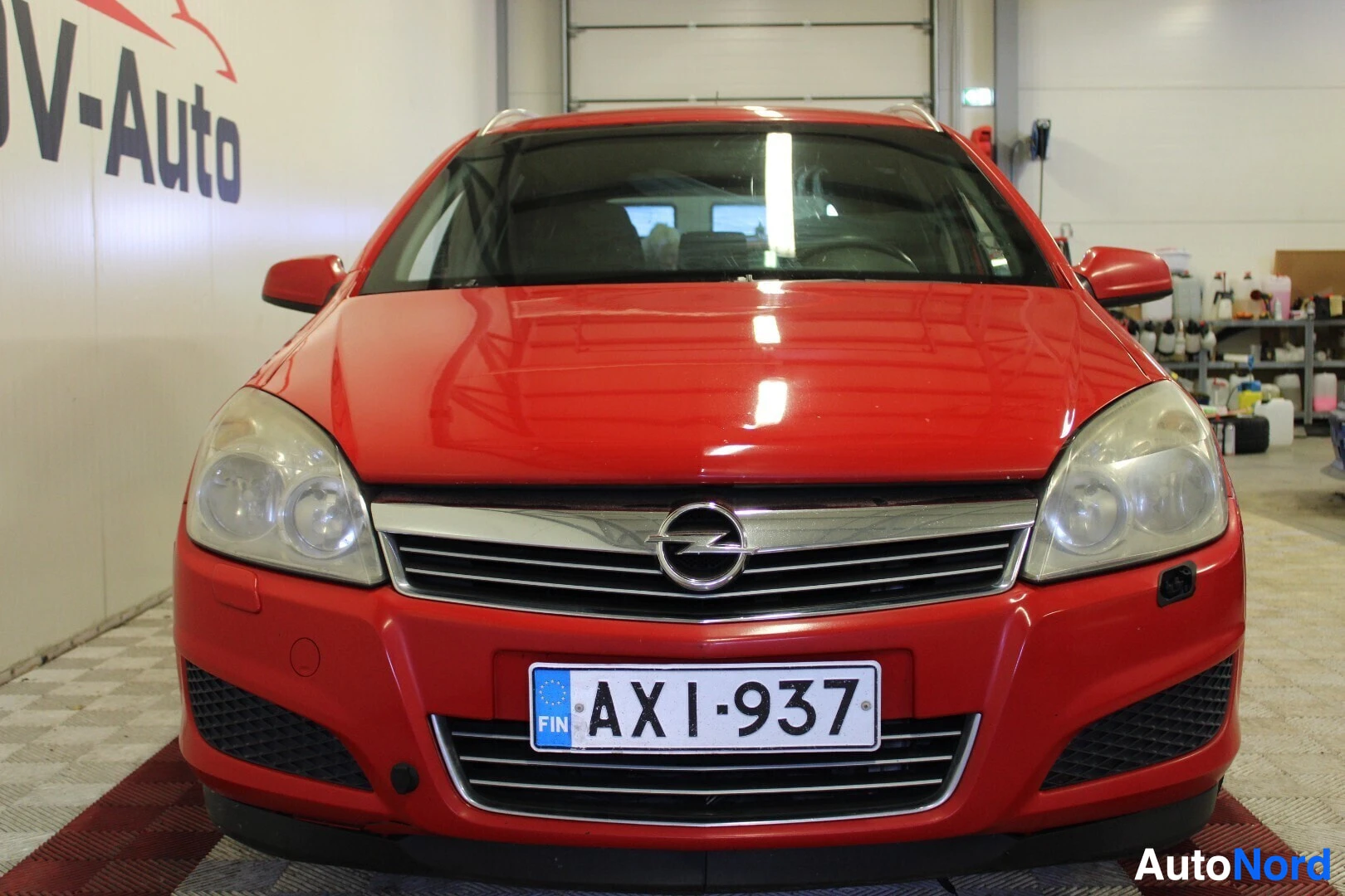Edestä: Punainen henkilöauto edestä päin kuvattuna. Etusäleikössä on hopeanvärinen poikkipalkki ja Opel-logo. Rekisterikilpi "FIN " on kiinnitetty etupuskuriin