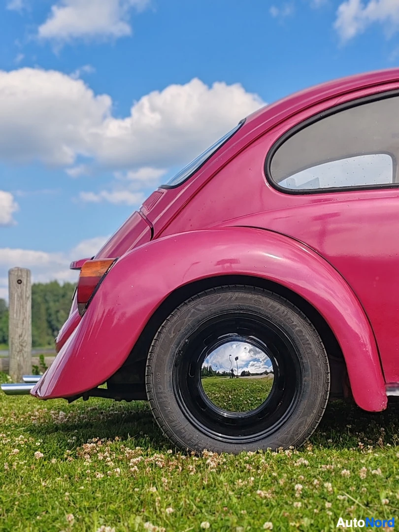 Yksityiskohta: Lähikuva vaaleanpunaisen Volkswagen Kuplan takaosasta ja mustasta vanteesta, jossa on kromattu pölykapseli. Taivaalla on pilviä, jotka heijastuvat pölykapselist