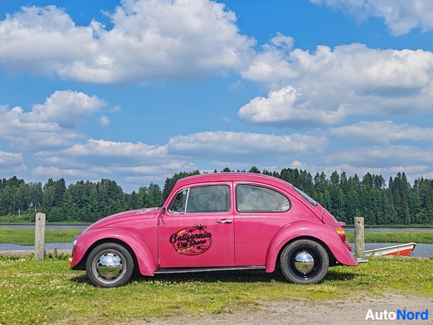 Sivulta: Vaaleanpunainen Volkswagen Kupla on pysäköitynä rannan ruohikolle. Auton kyljessä on "California Old Skool" -tarra. Taustalla näkyy vesistöä ja puita, sekä sini