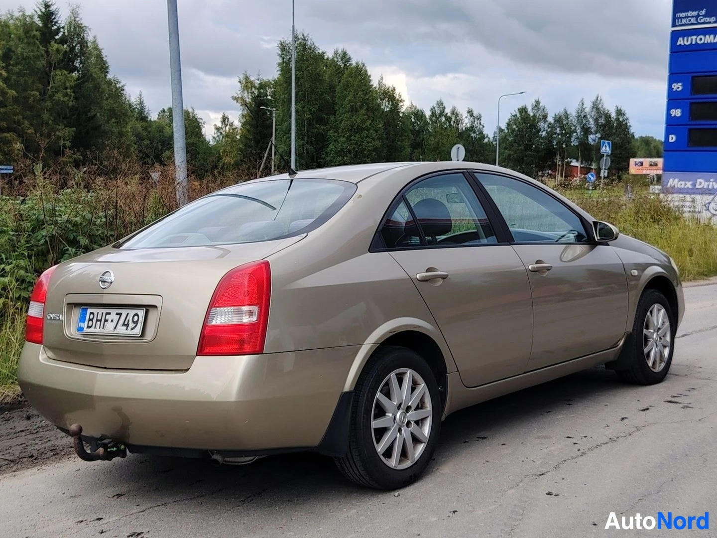 Takaviisto: Kultainen sedan-tyyppinen auto kuvattuna takaviistosta tien poskessa huoltoaseman kylttien ja metsätaustan edessä. Auton takaosa ja vasen kylki ovat näkyvissä.