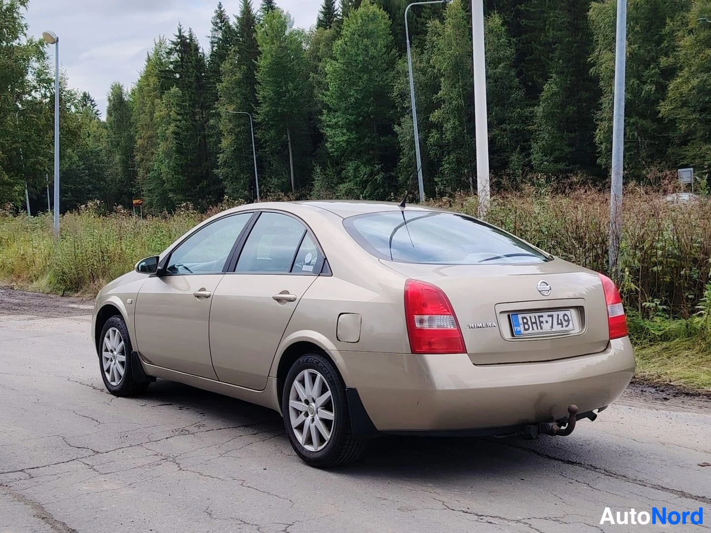 Takaviisto: Kultainen sedan-tyyppinen auto kuvattuna takaviistosta tien poskessa metsätaustan edessä. Auton takaosa ja vasen kylki ovat näkyvissä. Takarekisterikilpi näkyvi