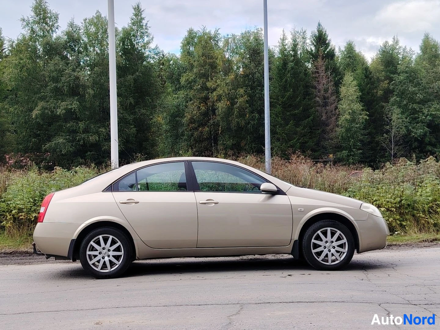 Sivulta: Beigen värinen sedan-tyyppinen henkilöauto on pysäköity sivuttain hiekkatielle metsätaustan edessä. Auton vasen kylki näkyy kokonaan.
