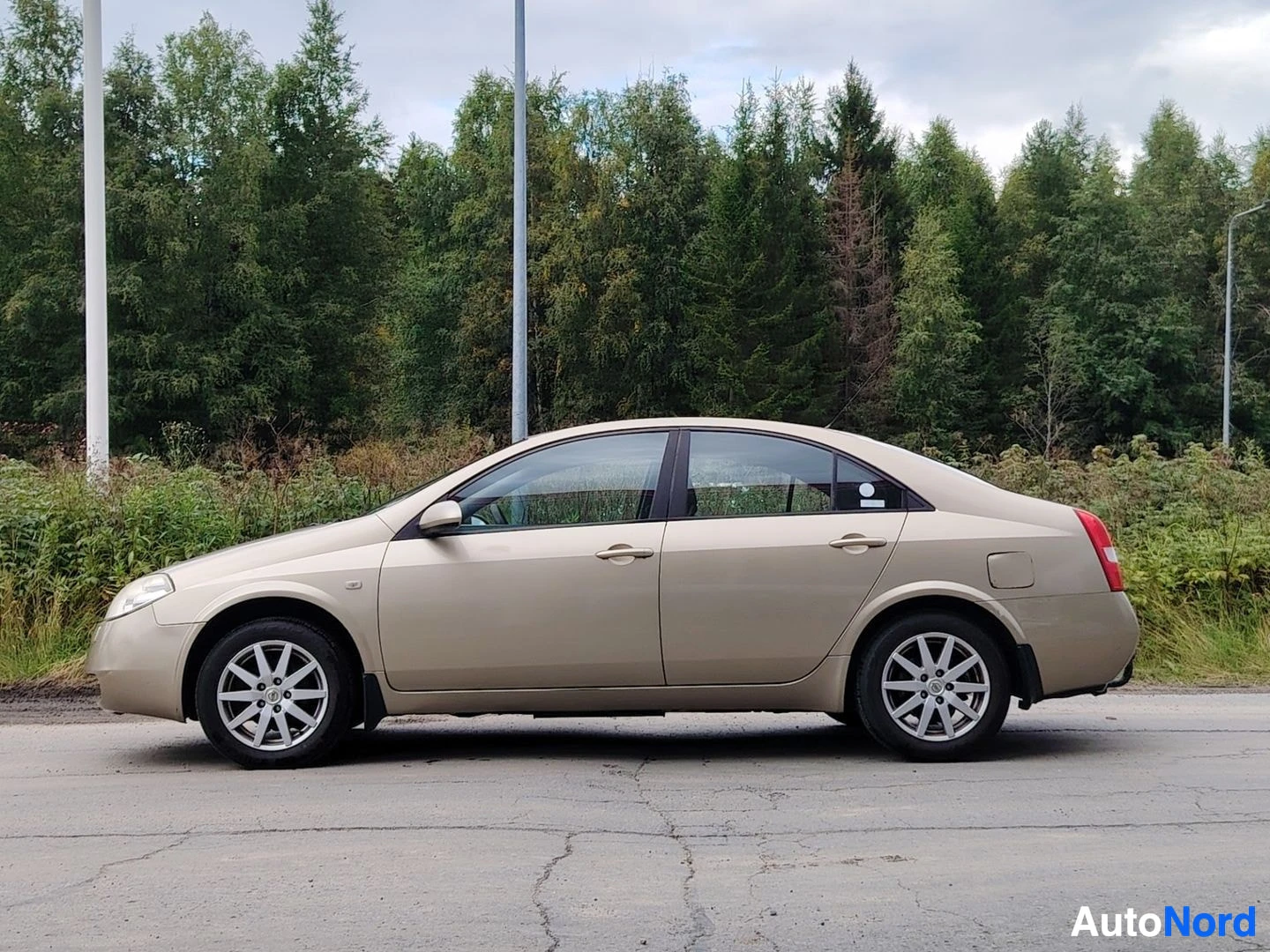 Sivulta: Beigen värinen sedan-tyyppinen henkilöauto on pysäköity sivuttain hiekkatielle metsätaustan edessä. Auton oikea kylki näkyy kokonaan.