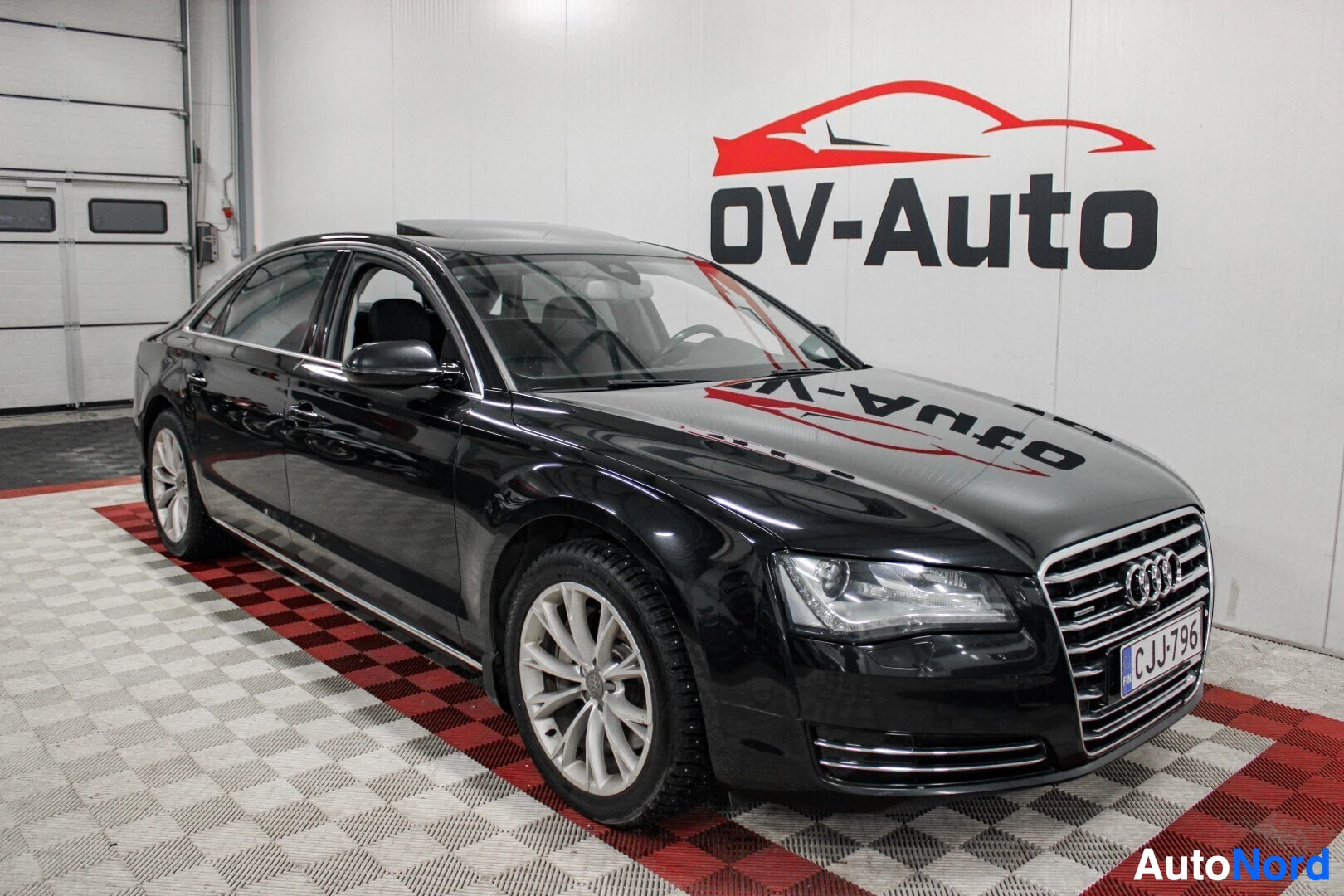 Etuviisto, nokka vasemmalle: Musta Audi A8 -henkilöauto pysäköitynä halliin, nähtynä edestä ja vasemmalta sivulta. Auton päällä on avattu kattoluukku. Ajoneuvon rekisterikilpi on "