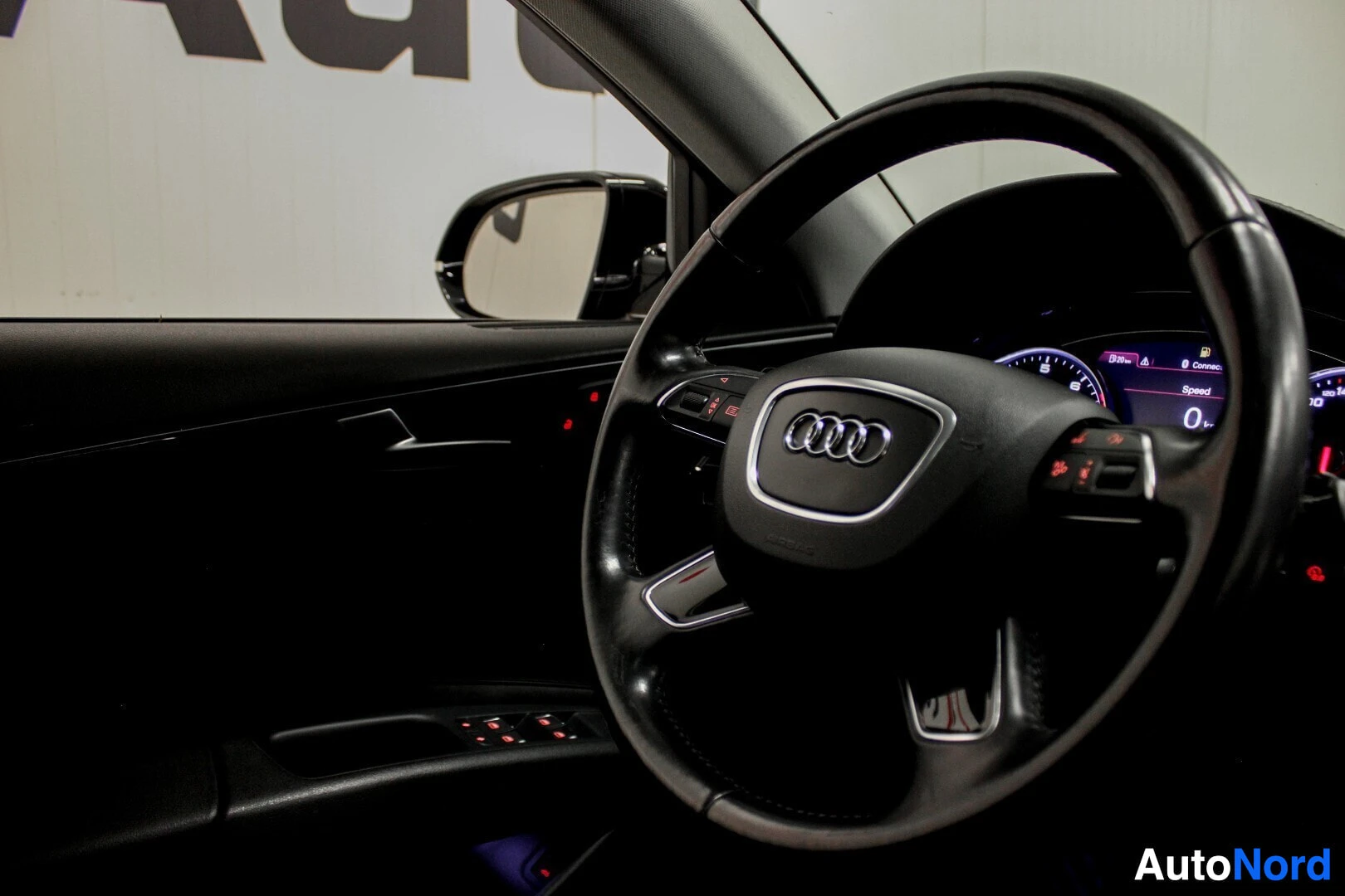 Yksityiskohta: Nahkainen ohjauspyörä, jossa on Audi-logo keskellä ja monitoimipainikkeita vasemmalla puolella. Kynnyslistassa ja ovessa on valoja. Peili heijastaa ulkopuolen.