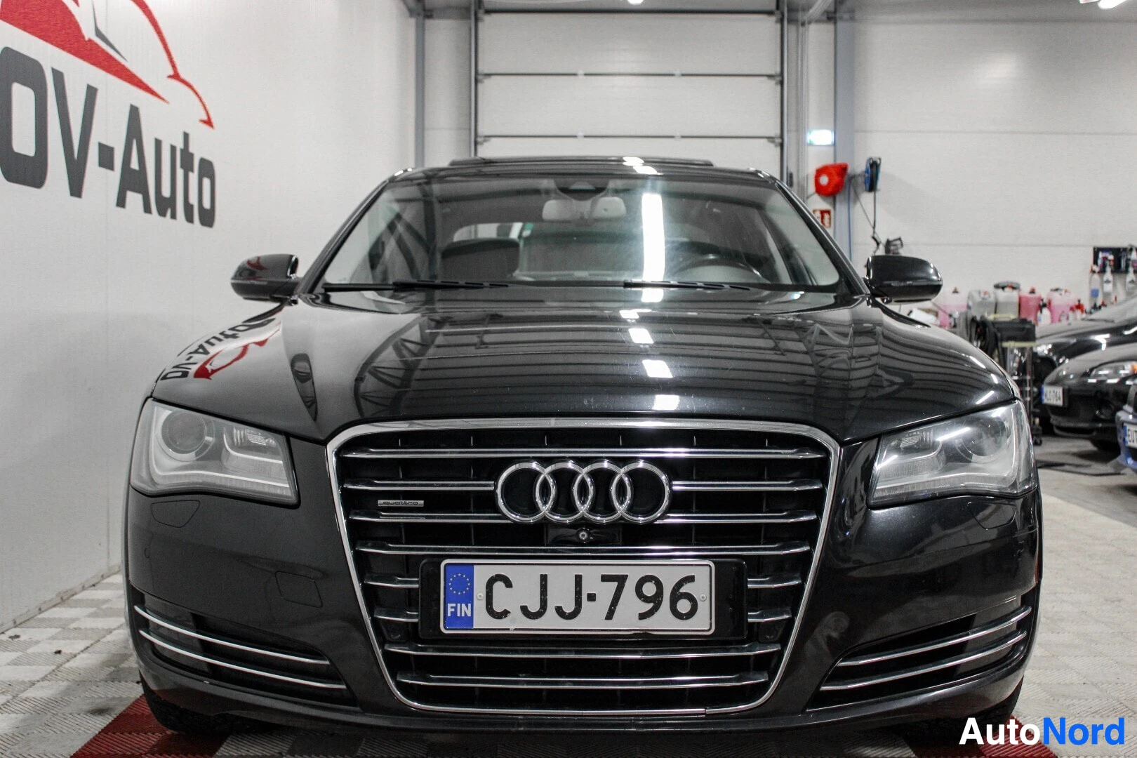 Edestä: Musta Audi A8 -henkilöauto edestä päin kuvattuna hallissa. Rekisterikilpi on ""