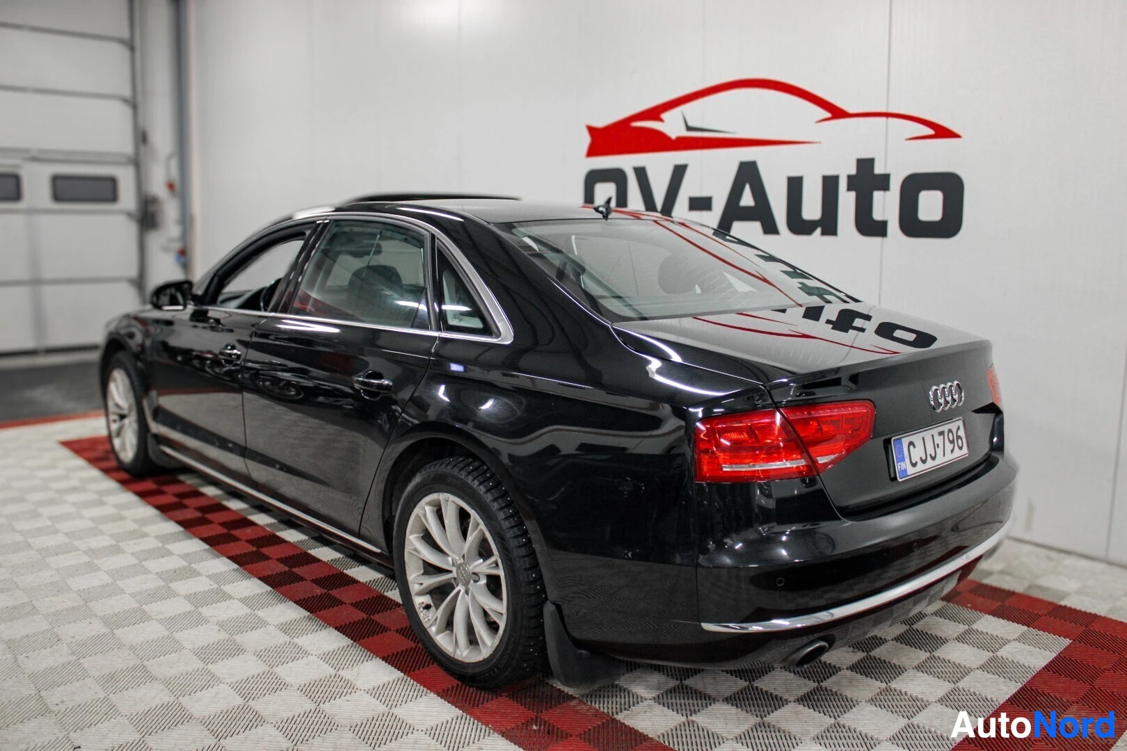 Takaviisto: Musta Audi A8 -henkilöauto pysäköitynä halliin, nähtynä takaa ja oikealta sivulta. Auton päällä on avattu kattoluukku. Ajoneuvon rekisterikilpi on ""