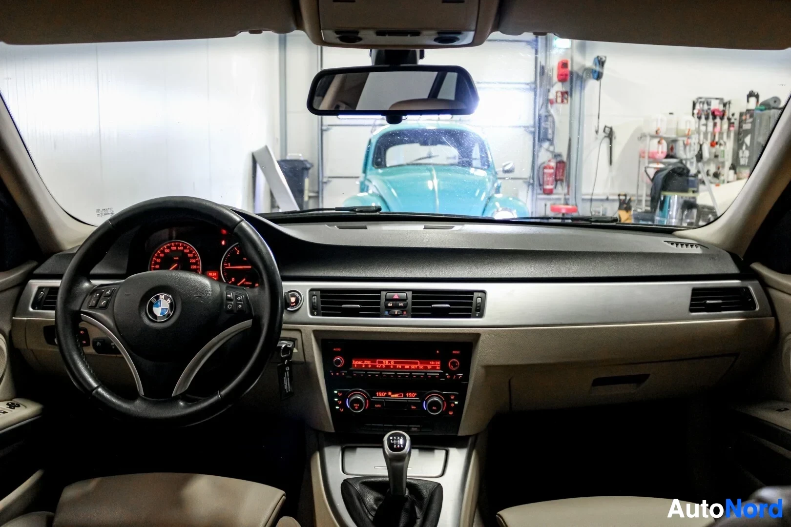 Ohjaamo: Sisäkuva auton ohjaamosta. Kojelauta, ohjauspyörä ja keskikonsoli ovat näkyvissä. Tuulilasin läpi näkyy autotallin takaosa ja vaaleansininen vanha auto.