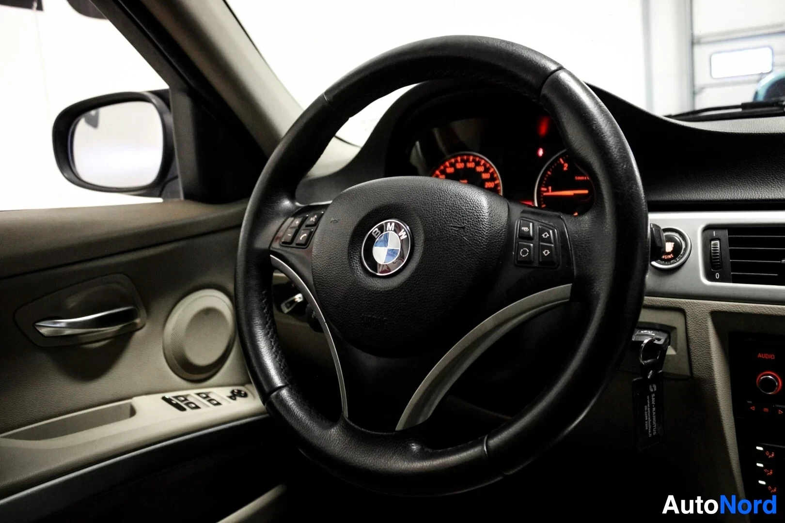 Yksityiskohta: Lähikuva auton ohjauspyörästä ja mittaristosta. Ohjauspyörässä on BMW-logo keskellä ja monitoiminäppäimet molemmilla puolilla. Mittariston taustavalo on punaine