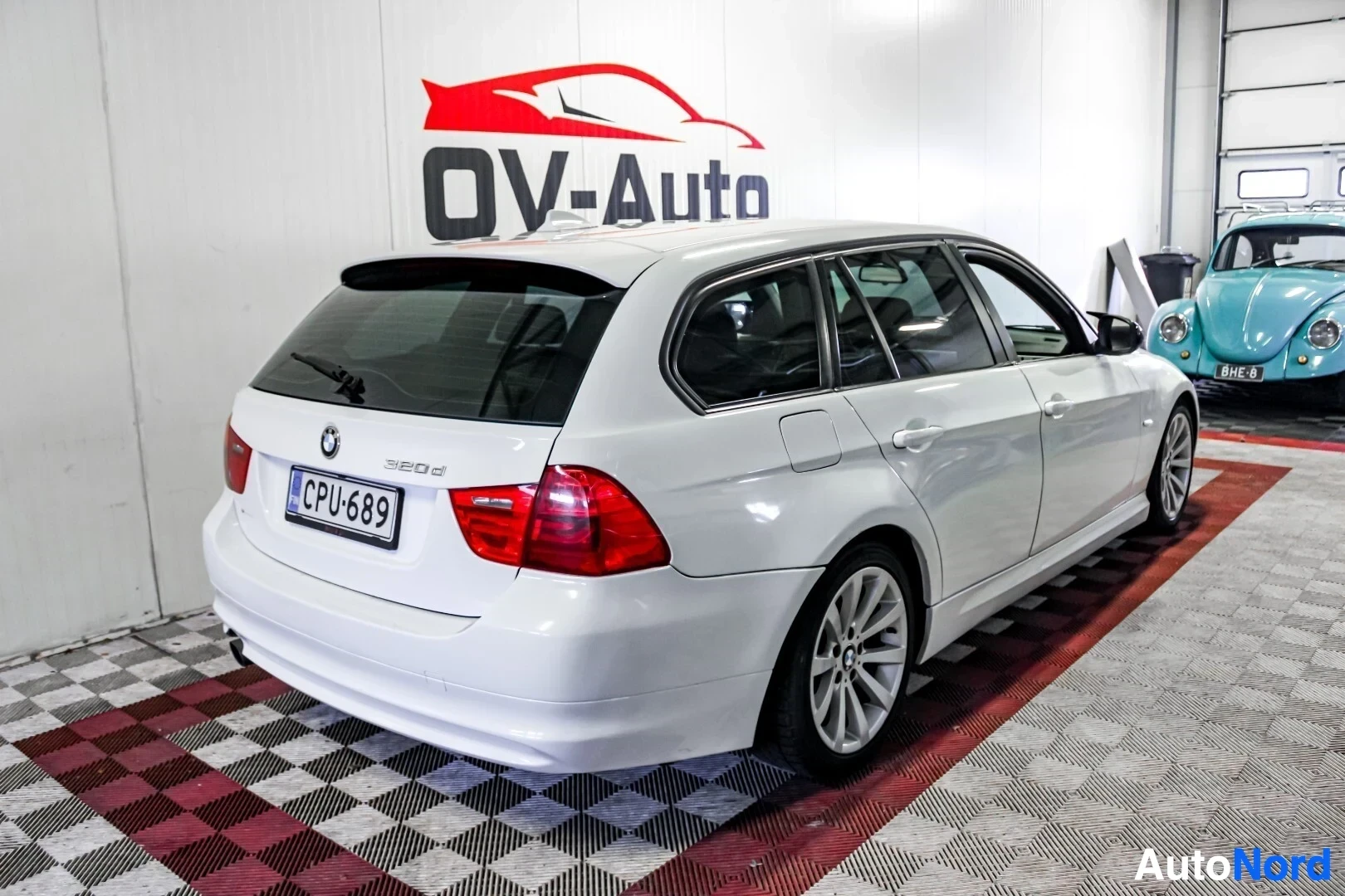 Takaviisto: Valkoinen BMW farmari on pysäköity sisätiloihin vasen takakulma kuvassa. Auton rekisterikilvessä lukee. Taustalla näkyy OV-Auto-logo ja osittain sininen