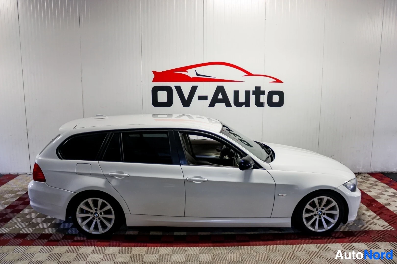 Sivulta: Valkoinen BMW farmari kuvattuna oikealta sivulta. Auto on pysäköity sisätiloihin shakkiruudulliselle lattialle, taustalla seinällä on OV-Auto-logo.