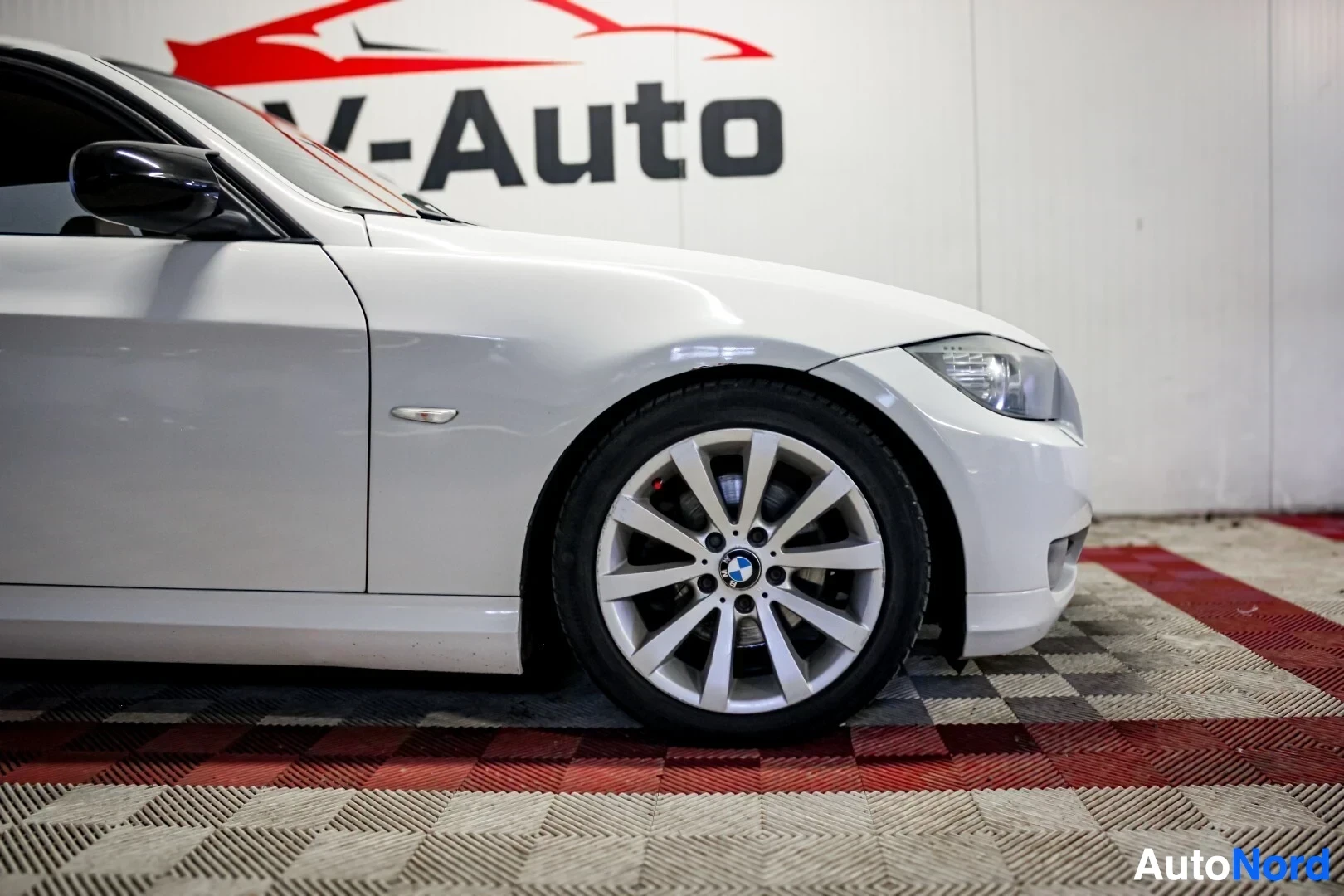 Yksityiskohta: Valkoisen BMW farmarin etupyörä ja osa etulokasuojaa ja ovea. Autossa on hopeanväriset kevytmetallivanteet. Taustalla näkyy OV-Auto-logo.