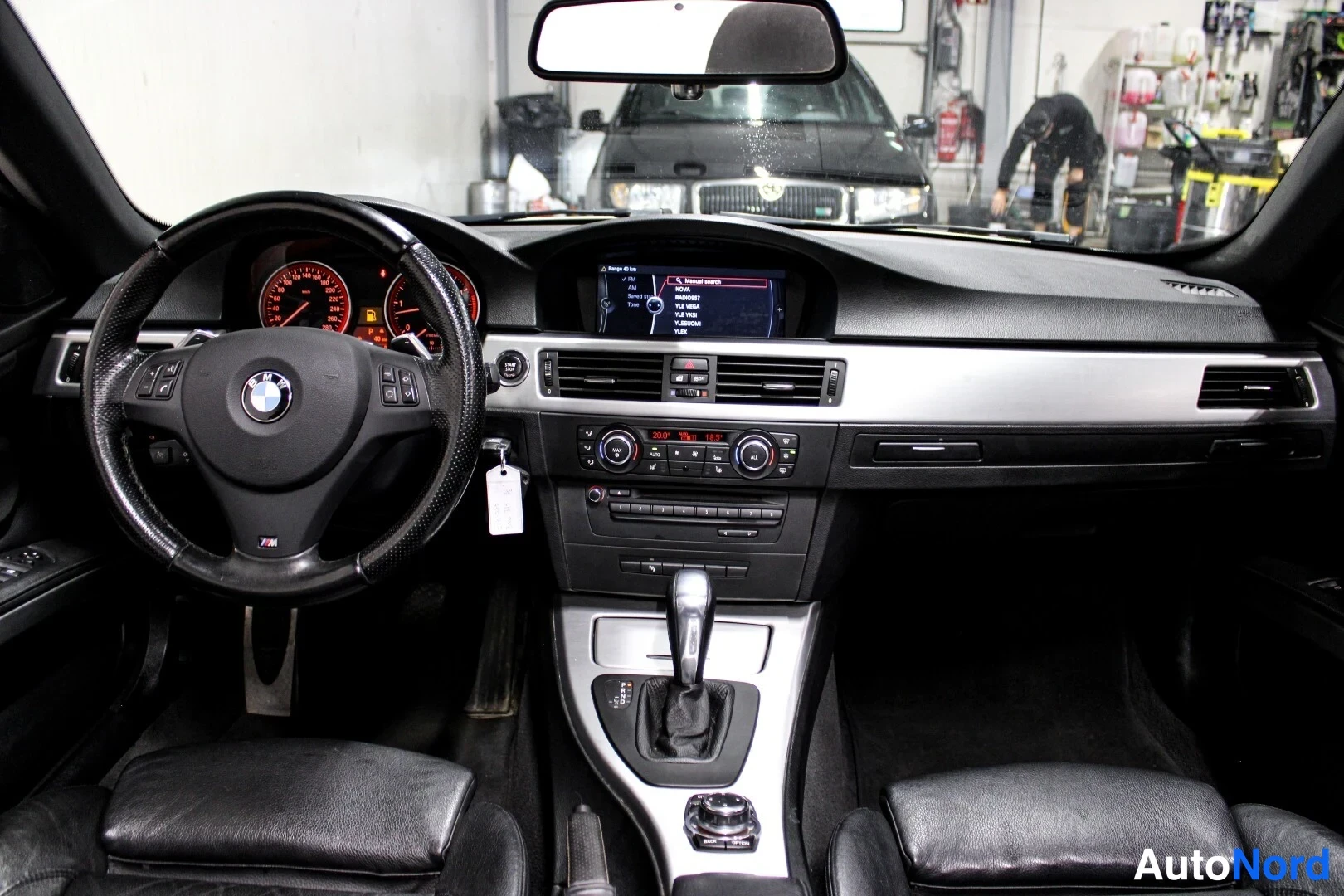 Muu: Näkymä etutuulasta: BMW:n mittaristo punaisilla valaisimilla, ruudulla menu, taustalla palokalusteita verstaalla.