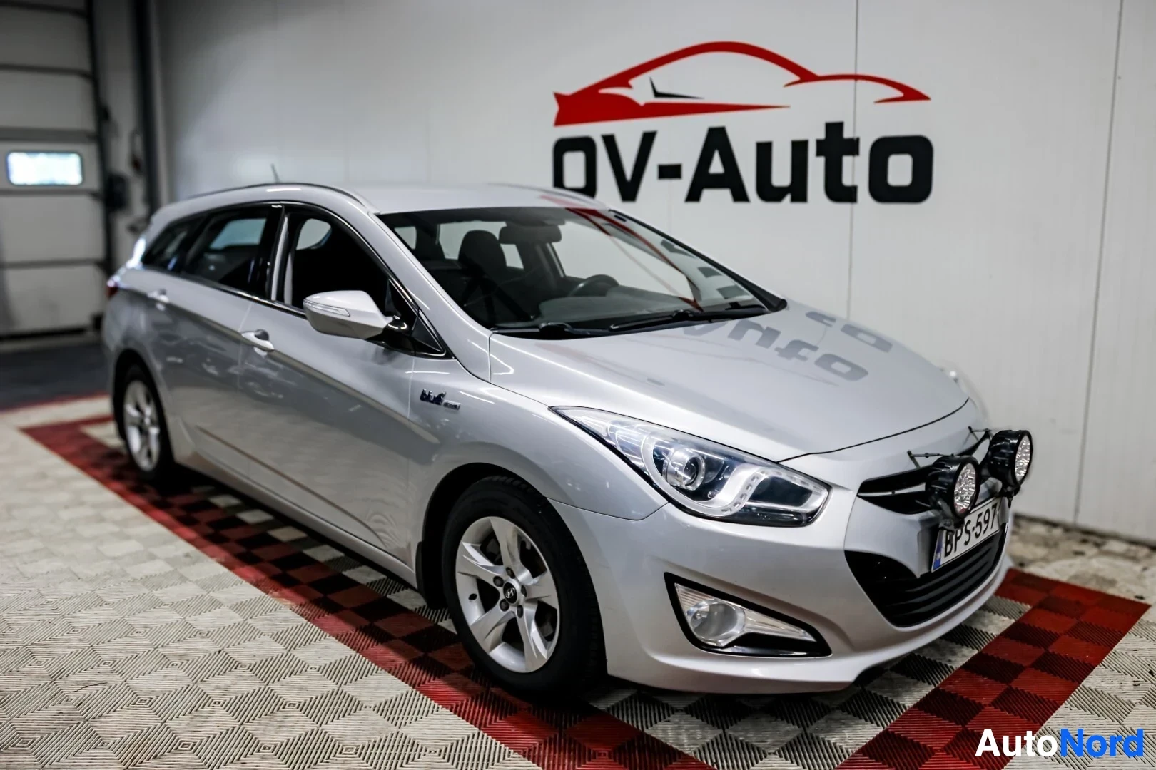 Etuviisto, nokka vasemmalle: Hopeanvärinen Hyundai i40 -farmariauto pysäköitynä sisätiloissa, etuvasemmalta kuvattuna. Autossa on lisävalot etupuskurissa ja kattokiskot katolla. Re