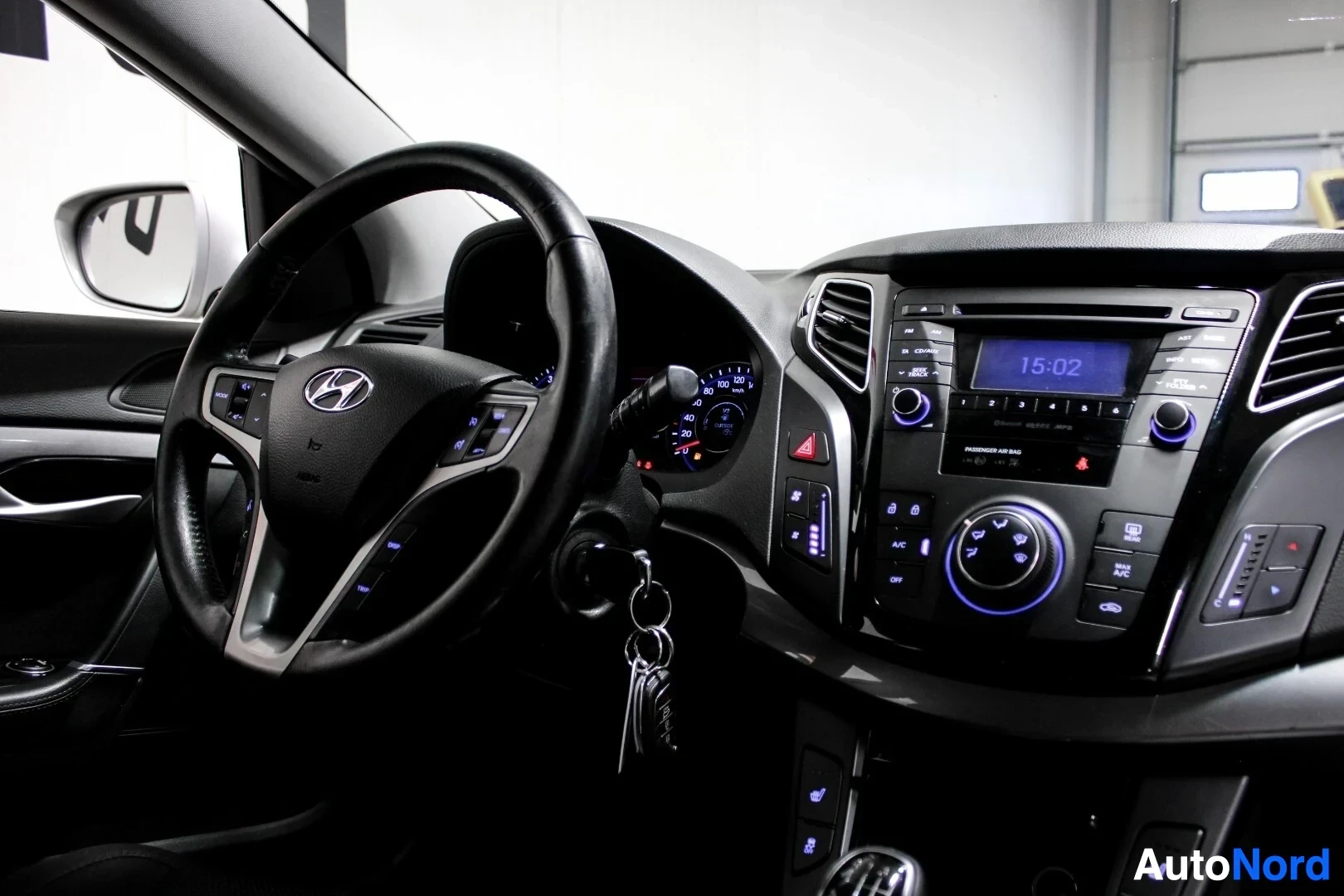 Ohjaamo: Kuvakulma oikealta edestä näyttää ohjaamon kojelaudan, ohjauspyörän Hyundai-logolla ja keskikonsolin painikkeet sekä näytön, jossa näkyy kello '15:02'. Ohjauspy