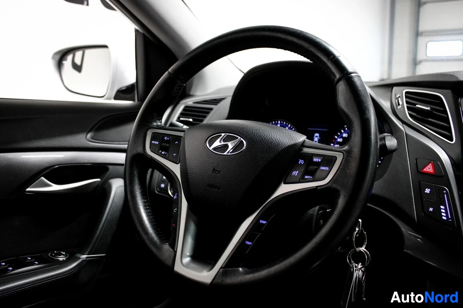 Yksityiskohta: Lähikuva ohjauspyörästä, jossa on Hyundai-logo ja useita painikkeita. Mittaristosta näkyy kirkkaita valoja. Ohjauspyörään kiinnitetyssä avainnipussa on kolme av