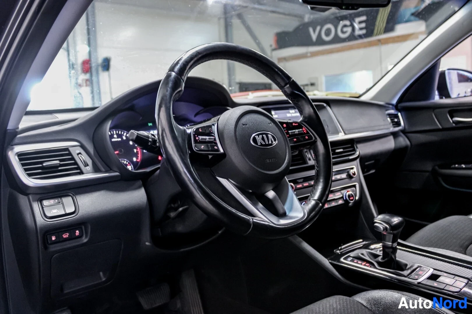 Ohjaamo: Kuvakulma auton ohjaamosta, joka näyttää ohjauspyörän, mittariston ja osan kojelautaa. Ohjauspyörässä on Kia-logo ja monitoimipainikkeita.