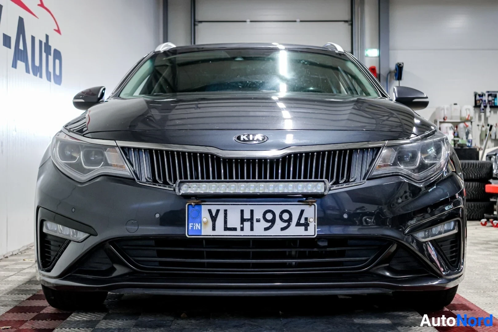 Edestä: Kia Optima farmariauton etuosa on kuvattu suoraan edestä sisätiloissa. Autossa on valkoiset rekisterikilvet ja puskurin yläpuolella lisävalo.