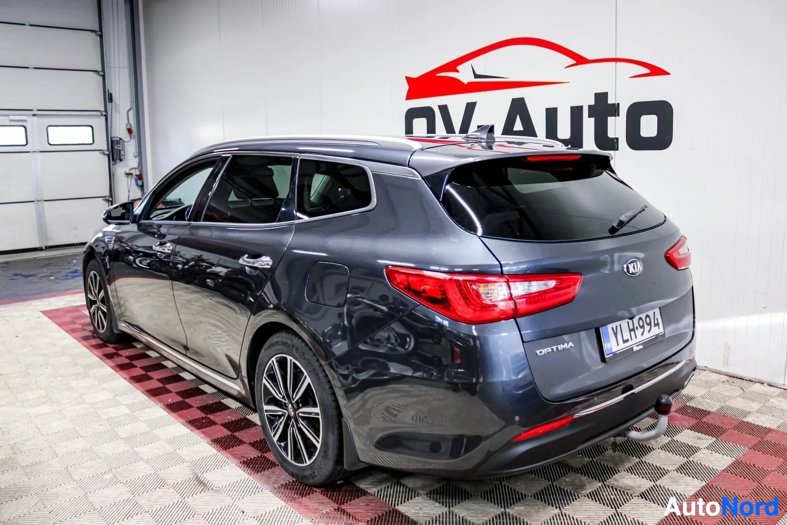 Takaviisto: Harmaa Kia Optima farmariauto on kuvattu takaa oikealta viistosta sisätiloissa, sen perä osoittaa kuvassa vasemmalle. Autossa on valkoiset rekisterikilvet ja ve