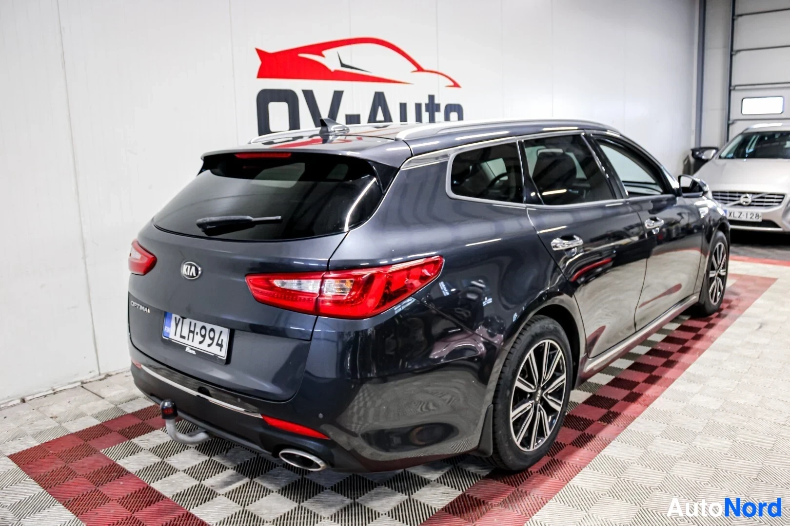 Takaviisto: Tummansininen Kia Optima farmari kuvattuna takavasemmalta viistosti. Auton takapää ja vasen kylki ovat näkyvissä. Autossa on vetokoukku, kattokaiteet ja kevytme