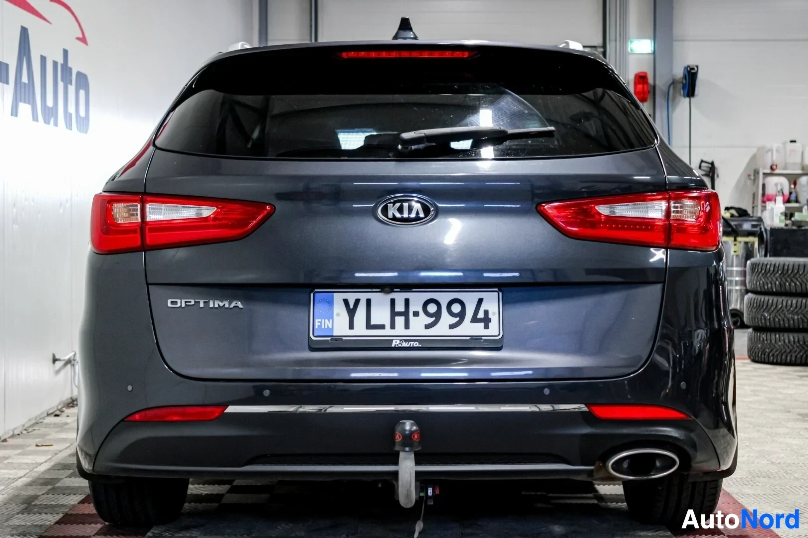 Takaa: Tummansininen Kia Optima farmari kuvattuna suoraan takaa. Auton takapäässä on vetokoukku ja yksi pakoputken ulostulo. Rekisterikilpi on näkyvissä. Kuvas