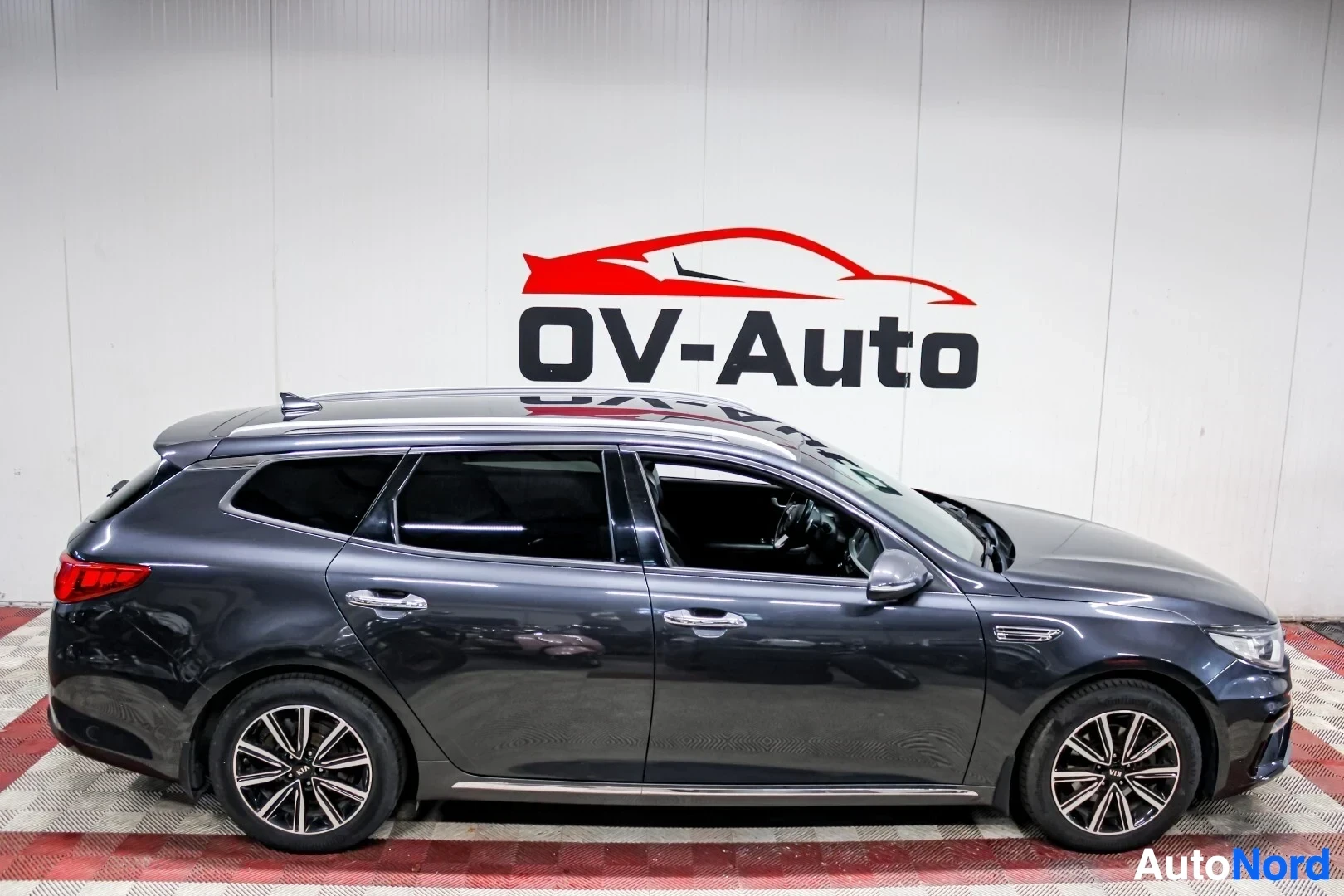 Sivulta: Tummansininen Kia Optima farmari kuvattuna vasemmalta sivulta. Autossa on kattokaiteet ja monipuolaiset kevytmetallivanteet. Koko ajoneuvo on näkyvissä.