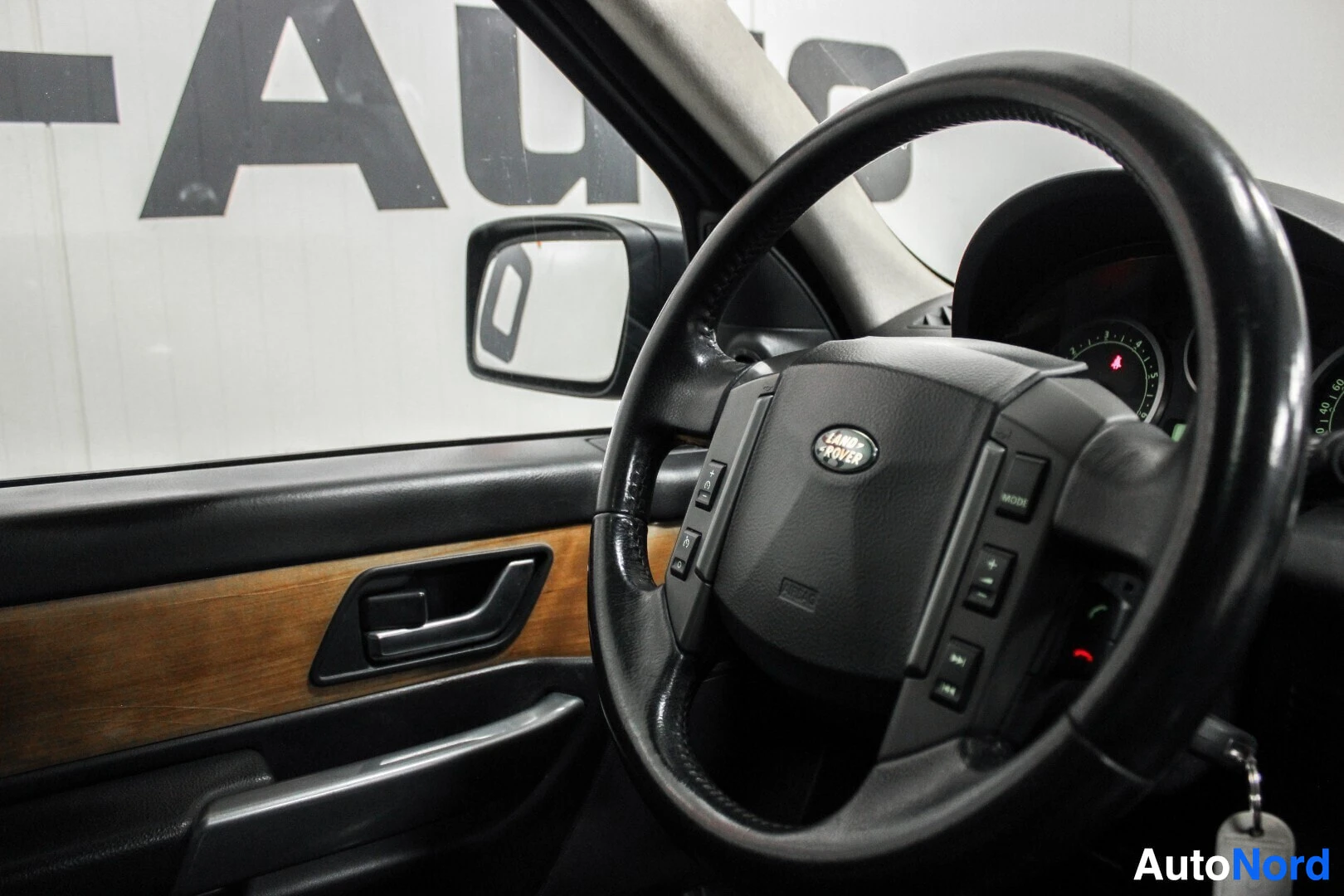 Ohjaamo: Kojelauta ja ohjauspyörä, jossa on painikkeet ja "LAND ROVER"-logo. Oikealla näkyy mittaristo ja vasemmalla puolella on musta ovipaneeli puunsyykuvioidulla osal