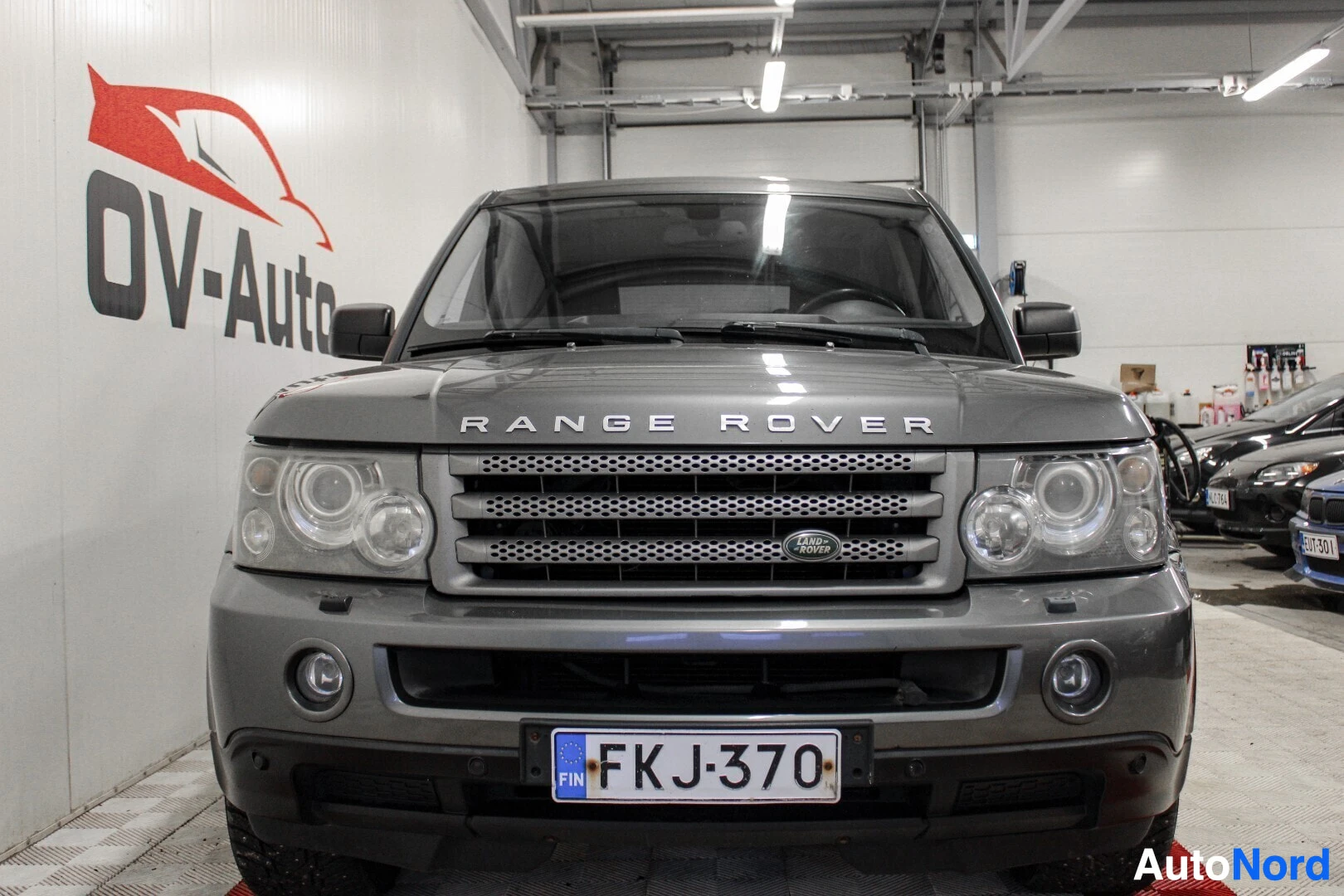 Edestä: Harmaa Land Rover Range Rover Sport -henkilöauto edestä päin kuvattuna. Autossa on "RANGE ROVER" -teksti konepellissä ja kromattu etusäleikkö, jossa on Land Rov