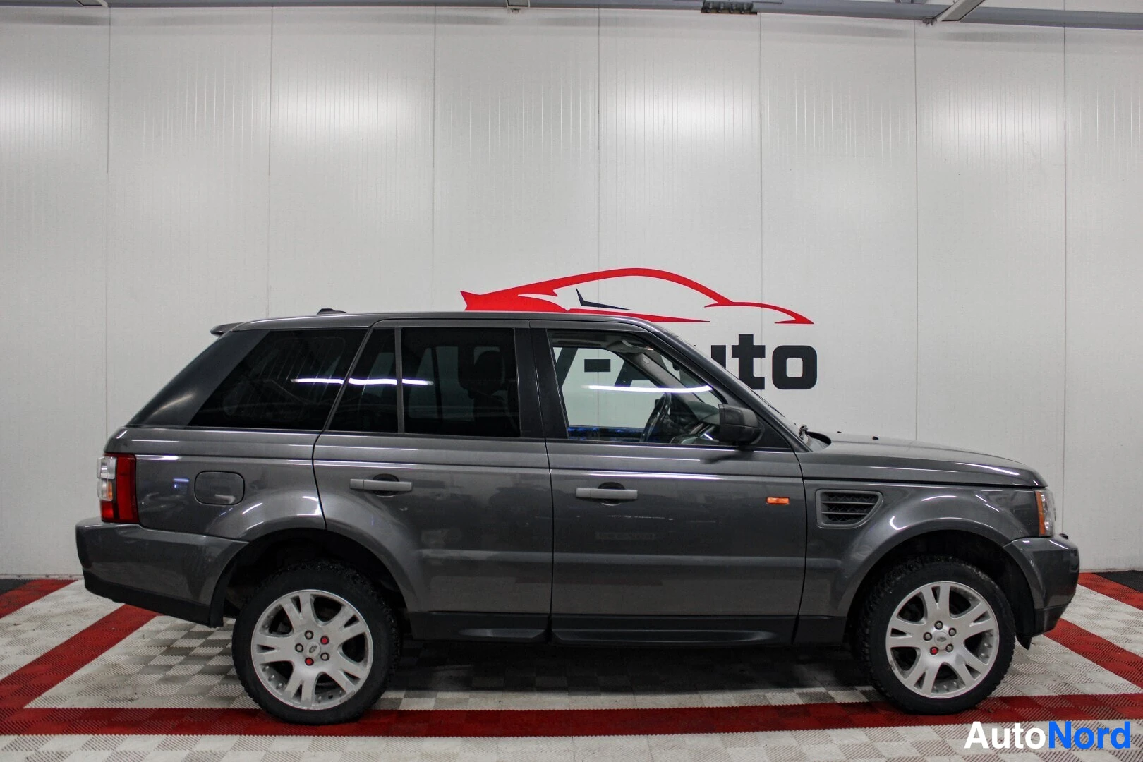 Sivulta: Harmaa Range Rover Sport vasemmalta sivulta sisätiloissa. Auto on parkissa punavalkoisella ruutulattialla.