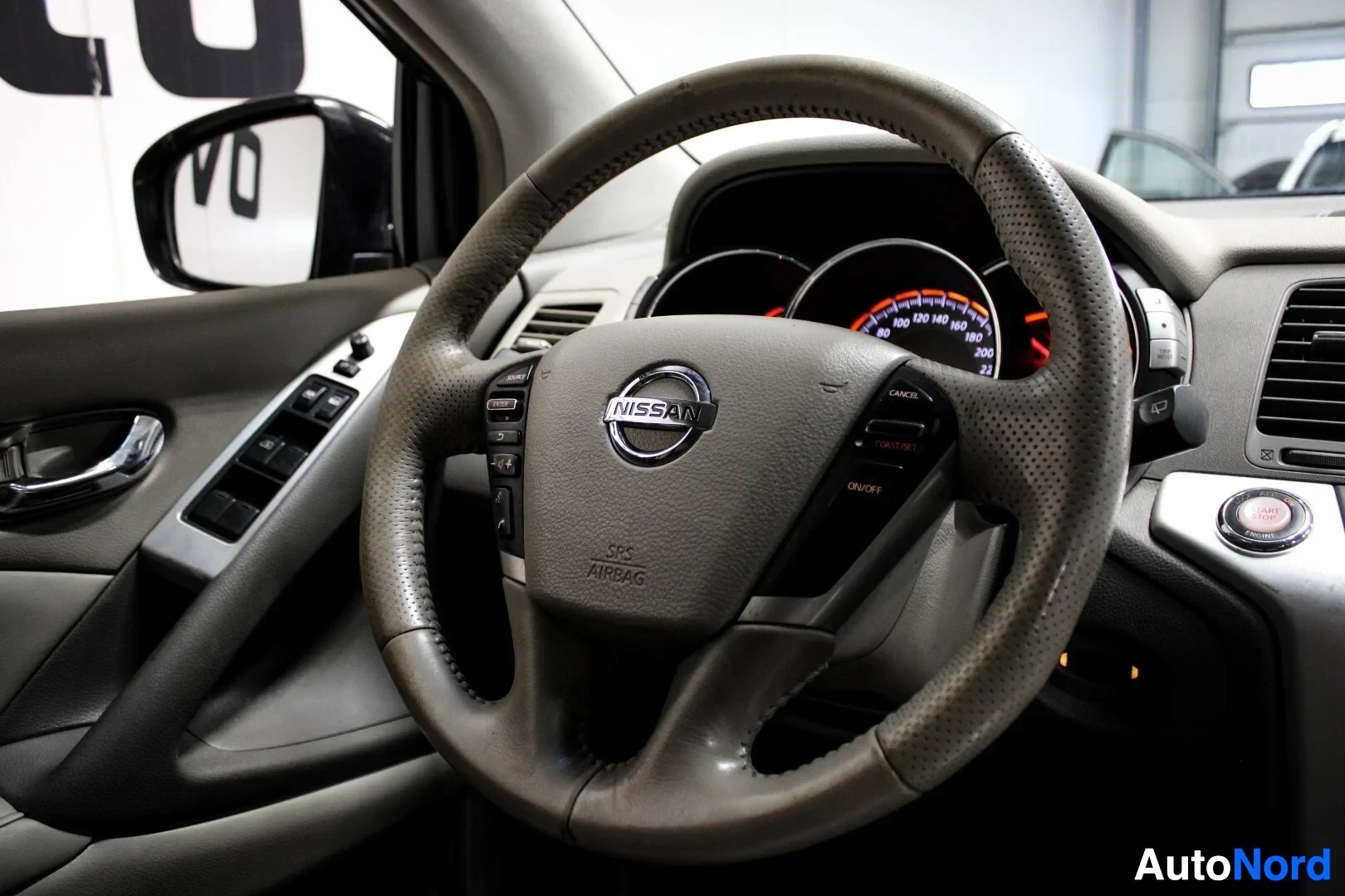 Yksityiskohta: Lähikuva ohjauspyörästä, jossa on Nissan-logo ja painikkeet rattipolkimilla sekä 'SRS AIRBAG' -merkintä.