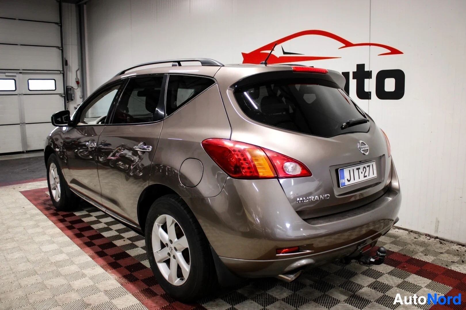 Takaviisto: Beige SUV takaa vasemmalta, pysäköitynä ruudulliselle lattialle sisätiloissa. Autossa on vetokoukku, kattokaiteet ja takaikkunan pyyhin. Taustalla näkyy yrityks