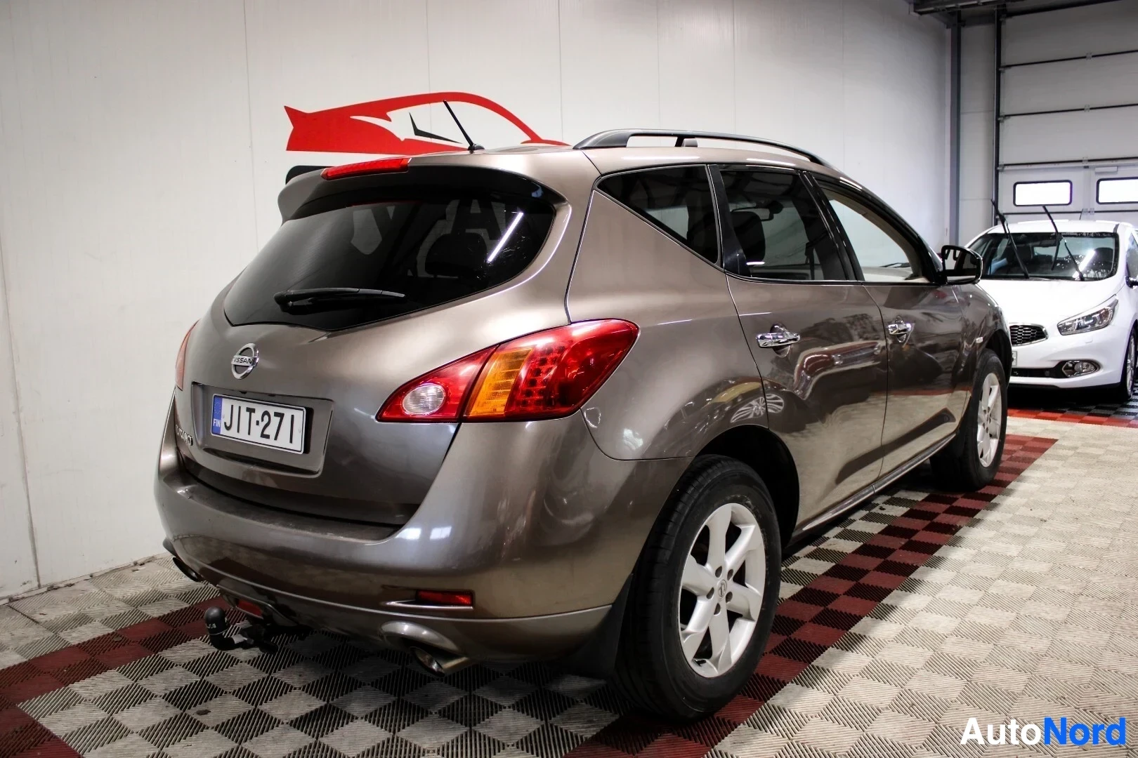 Takaviisto: Nissan Murano, jonka tummanruskea koripaneeli heijastaa valoa, nähtynä takaviistosta oikealta puolelta. Autossa on kattokaiteet ja vetokoukku.