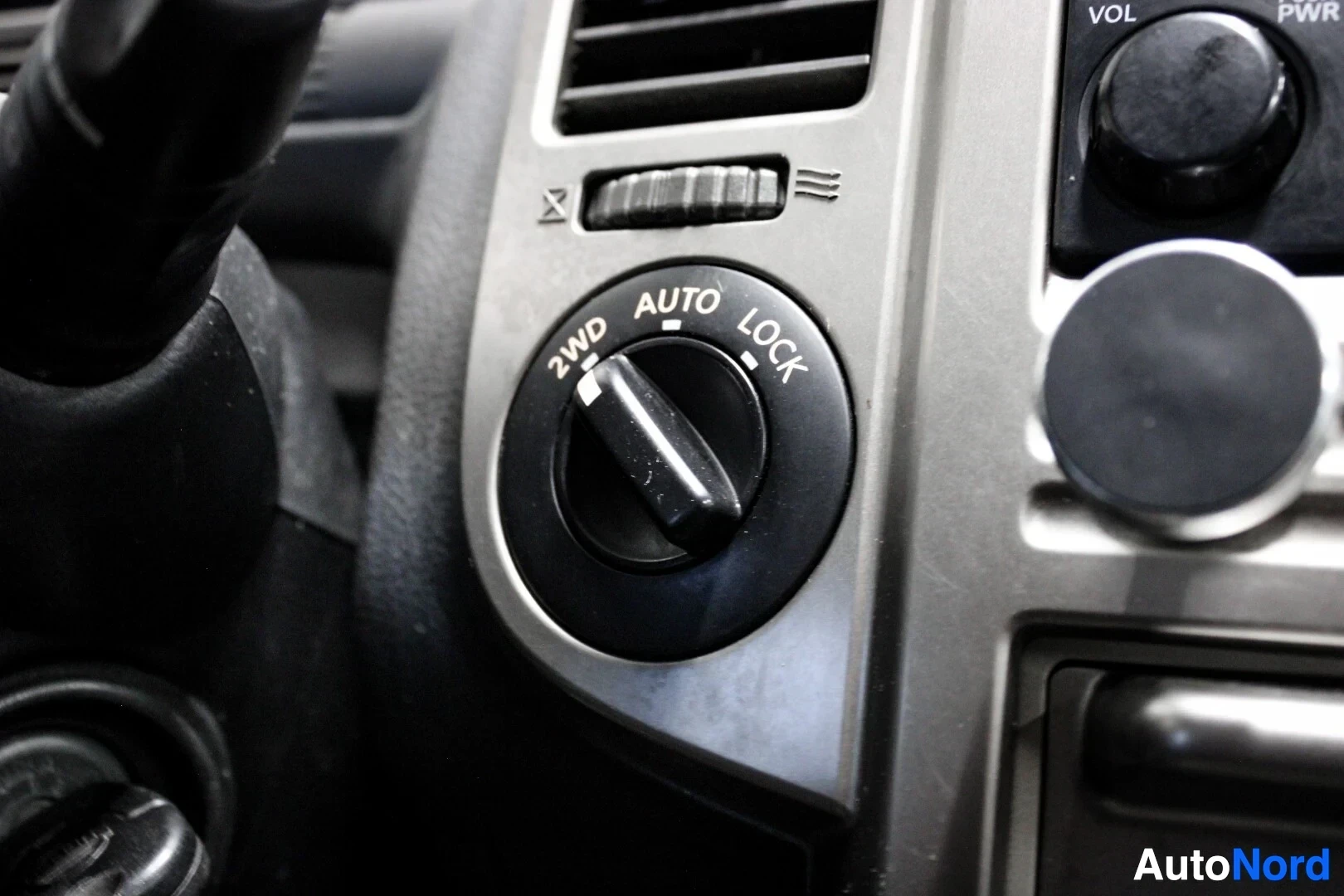 Ohjaamo: Lähikuva kojelaudan valitsimesta, jossa on asennot "2WD", "AUTO" ja "LOCK". Valitsin on asetettu "AUTO"-asentoon. Ilmastoinnin suulakkeet ja muut säätimet ovat