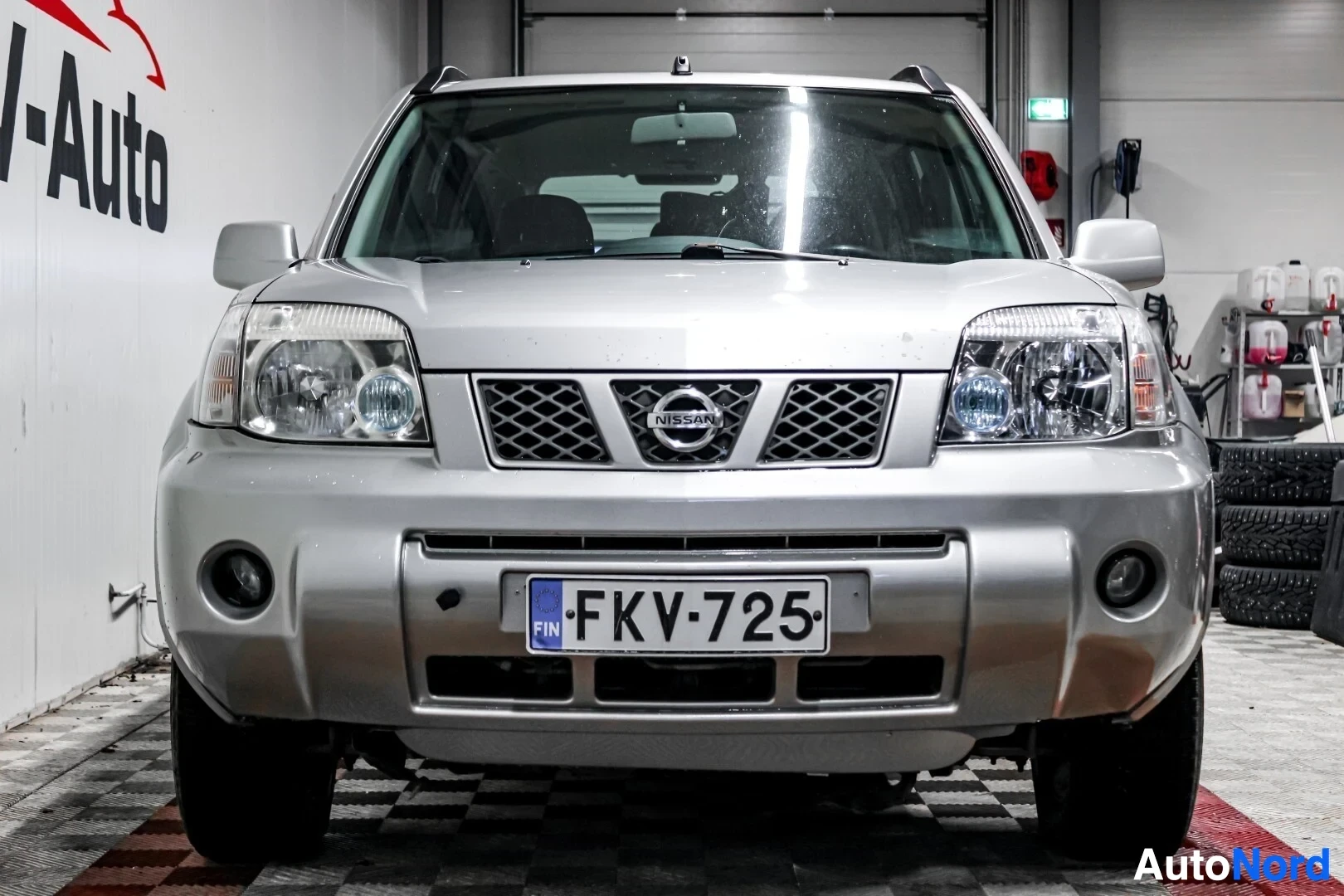 Edestä: Hopeanvärinen Nissan X-Trail edestä päin kuvattuna autohallissa. Autossa on rekisterikilpi "" ja etupuskurissa sumuvalot. Taustalla näkyy OV-Auto-logo ja
