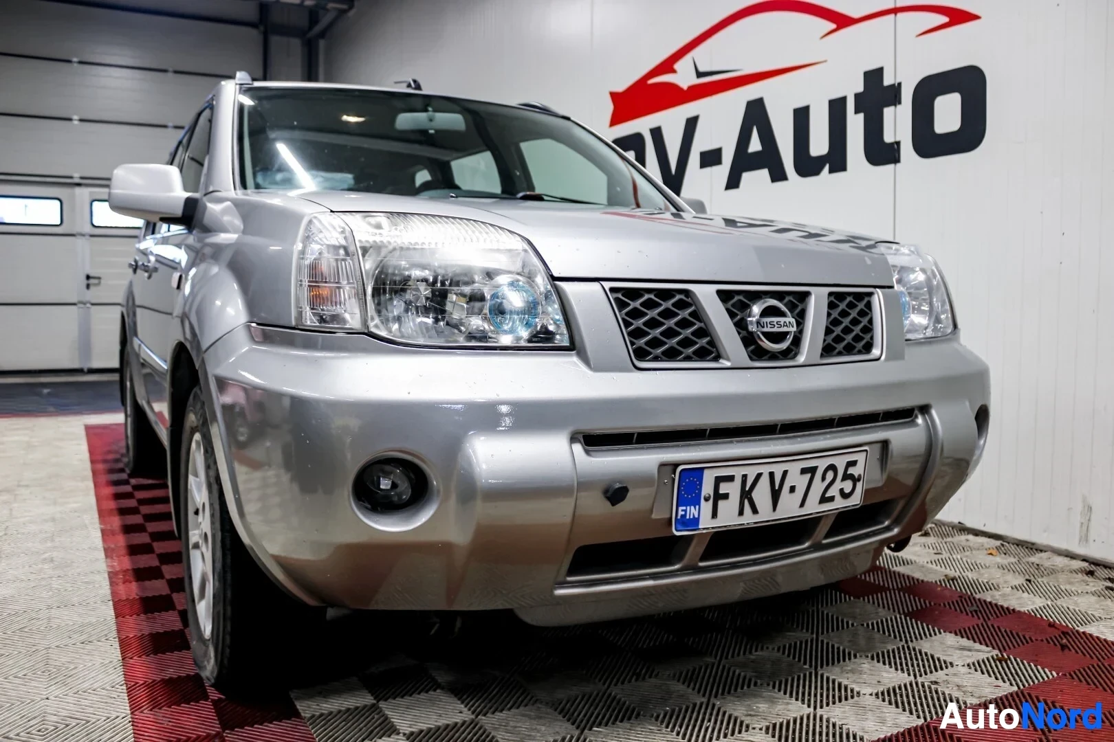 Etuviisto, nokka vasemmalle: Hopeanvärinen Nissan X-Trail edestä oikealta matalasta kulmasta kuvattuna. Rekisterikilvessä lukee "" ja auton etupuskurissa on sumuvalot