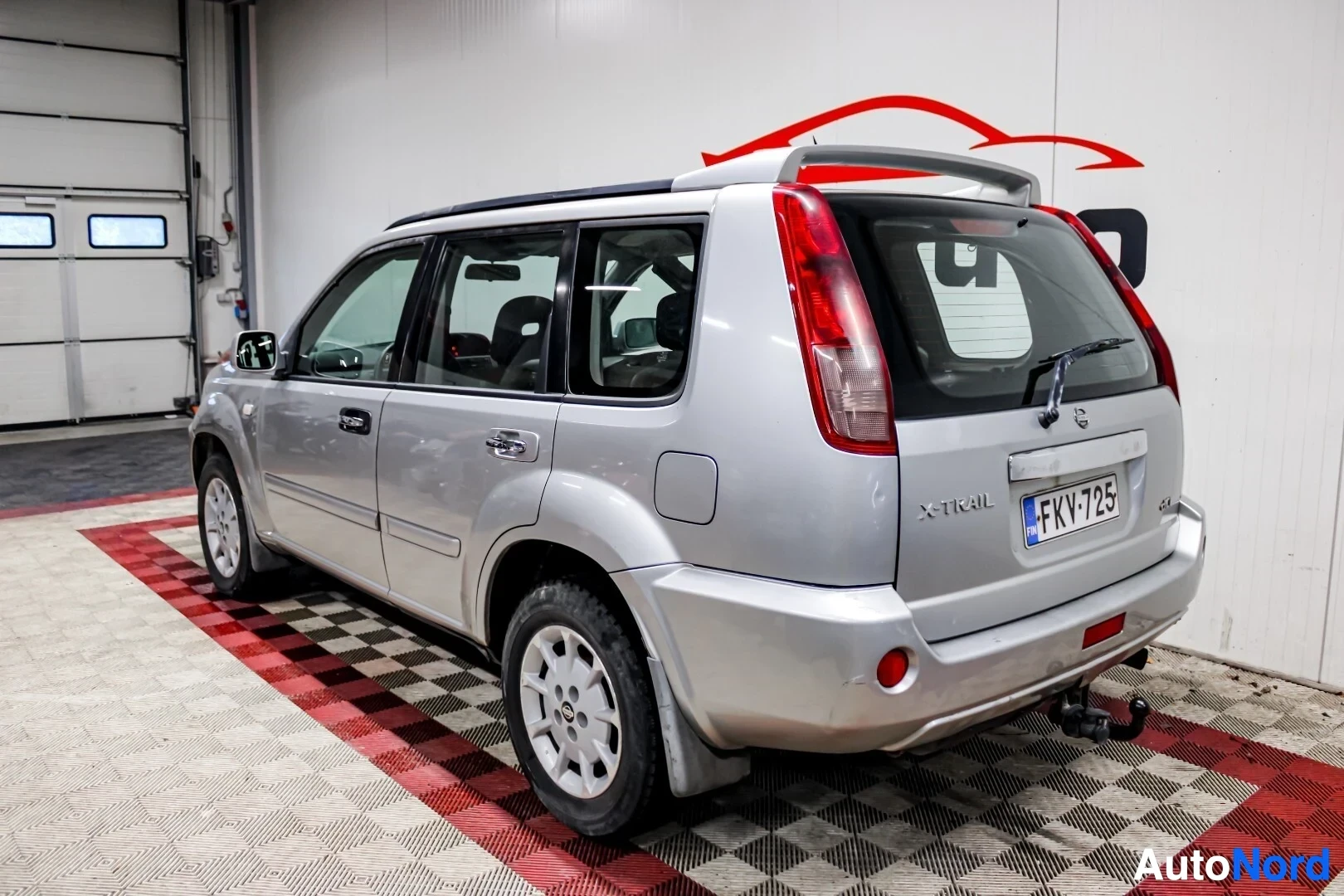 Takaviisto: Hopeanvärinen Nissan X-Trail takaviistosta oikealta kuvattuna sisätiloissa, takaluukun vasemmassa alakulmassa on "X-TRAIL"-merkintä. Auton perässä on vetokoukku