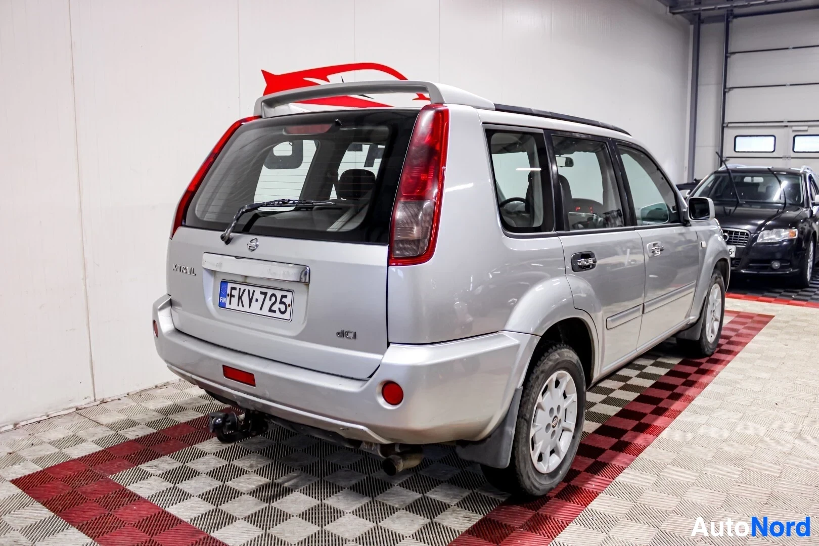 Takaviisto: Hopeanvärinen Nissan X-Trail takaviistosta vasemmalta kuvattuna sisätiloissa, takaluukun vasemmassa alakulmassa on "X-TRAIL"-merkintä ja oikeassa "dCi". Auton p
