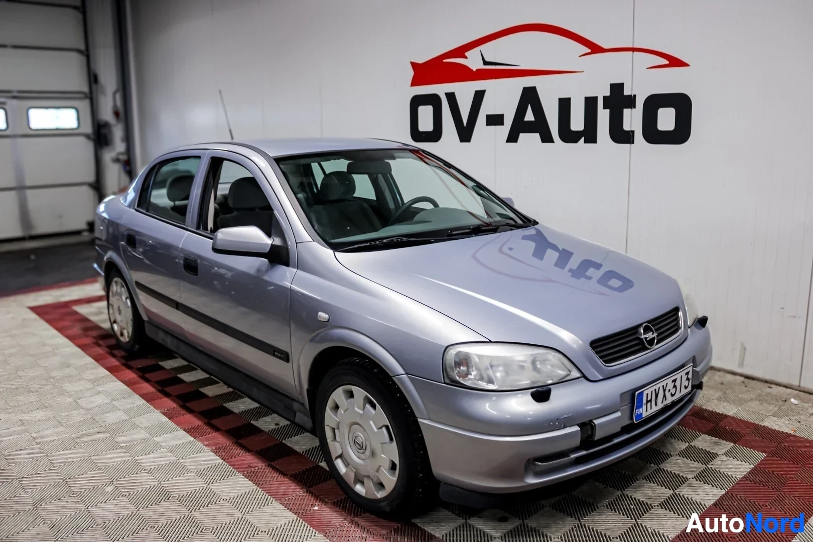 Etuviisto, nokka oikealle: Harmaa Opel Astra -henkilöauto, jossa on neljä ovea ja pyörissä pölykapselit, on kuvattuna sisältäpäin vasemmalta edestä, kohti auton oikeaa takaosaa. Au