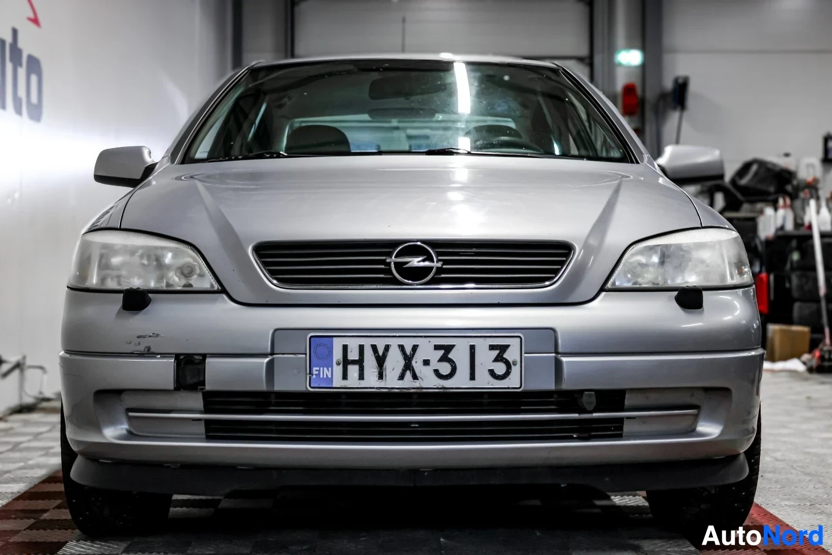 Edestä: Harmaan Opel Astra -henkilöauton keula edestä päin kuvattuna. Autossa on ajovalot ja maski, jonka keskellä on Opel-logo. Etupuskurissa on rekisterikilpi.