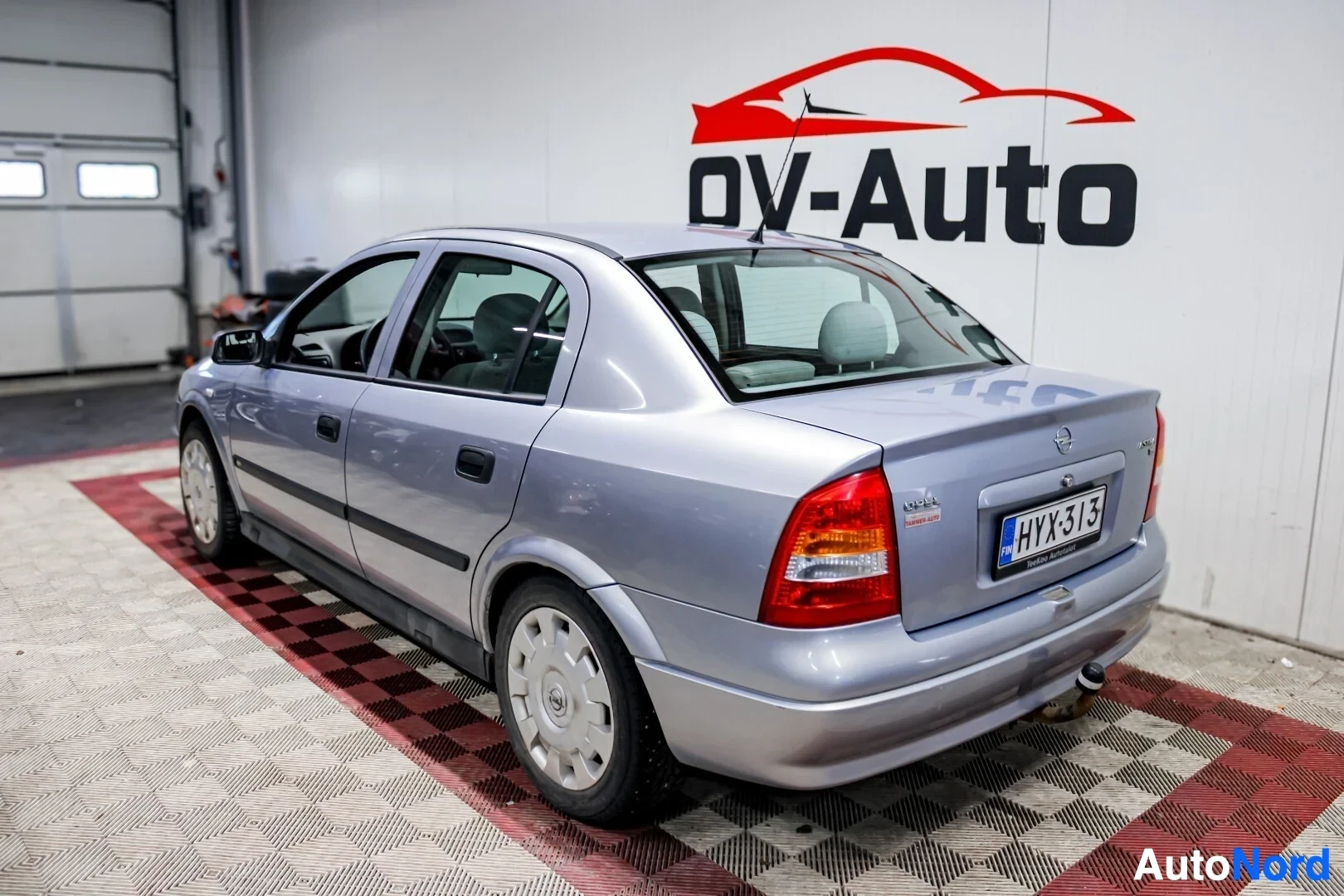 Takaviisto: Harmaa Opel Astra -henkilöauto, jossa on neljä ovea ja pyörissä pölykapselit, on kuvattuna takaa oikealta. Auton perä on kohti kuvan vasenta reunaa. Autossa on