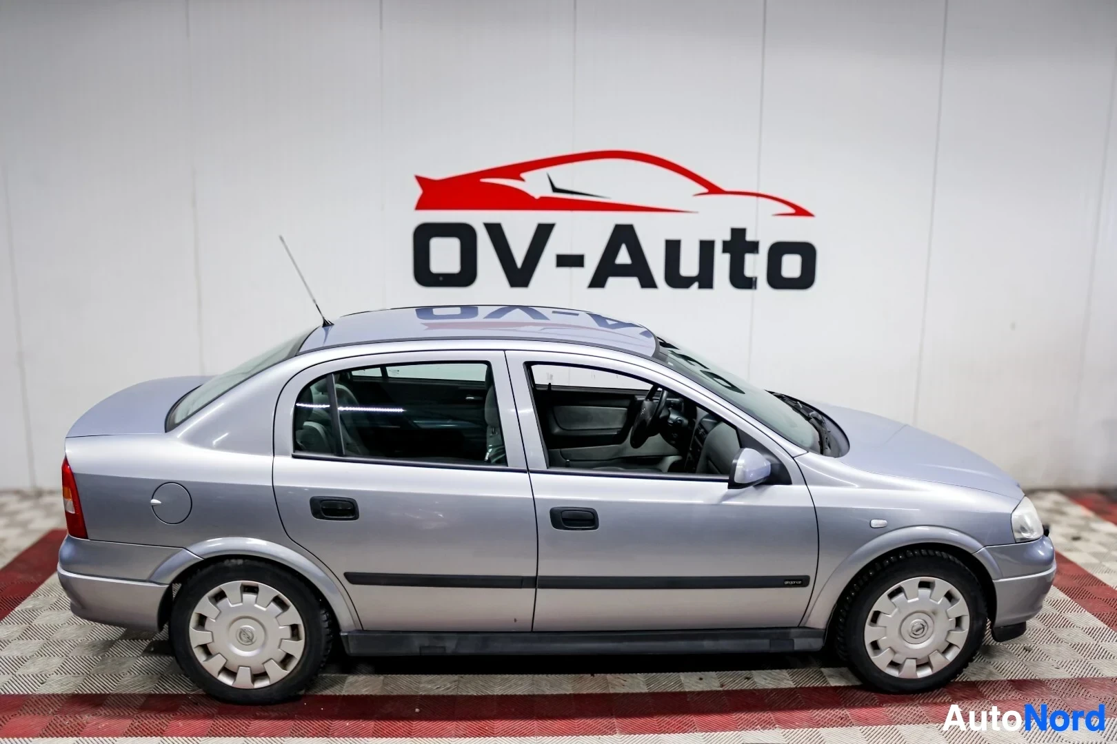 Sivulta: Hopeanvärinen Opel Astra sedan kuvattuna oikealta sivulta sisätiloissa. Autossa on kapselit vanteissa.