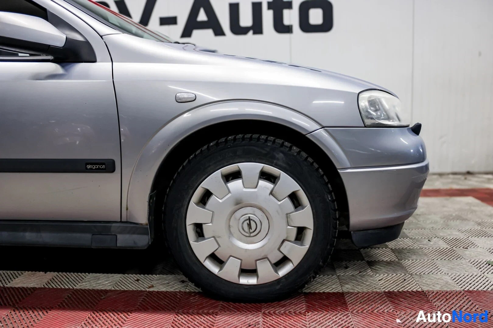 Yksityiskohta: Lähikuva hopeanvärisen Opel Astran oikeasta etupyörästä ja etulokasuojasta. Lokasuojassa lukee 'elegance'.