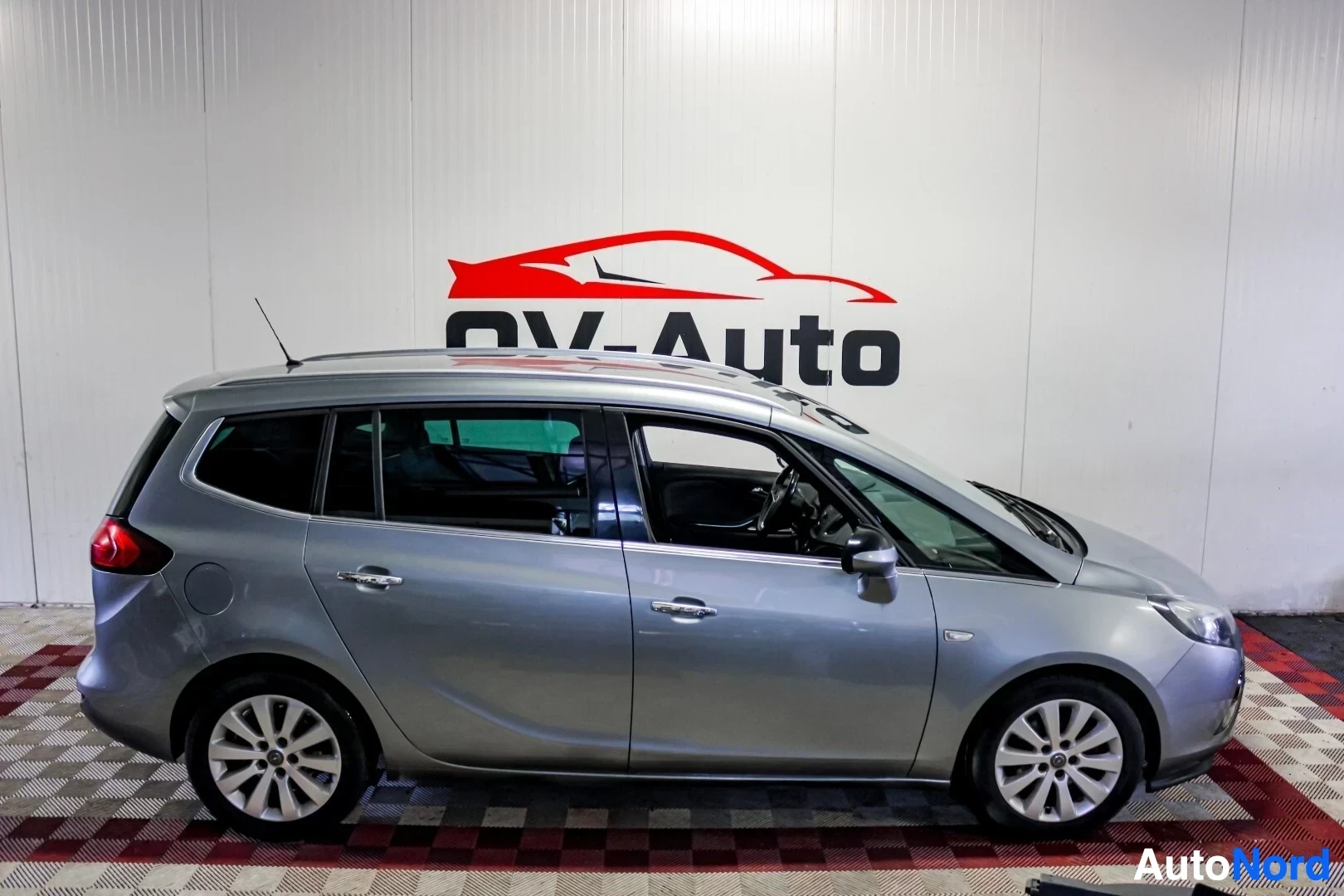 Sivulta: Hopeanvärinen tila-auto oikealta sivulta kuvattuna valkoisen seinän edessä.