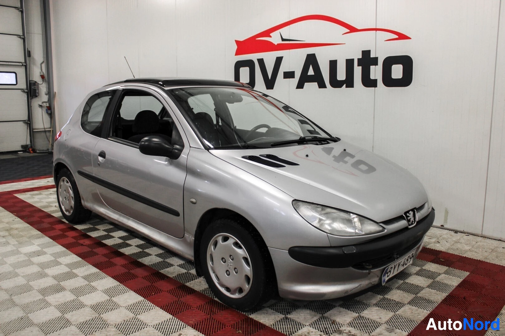 Etuviisto, nokka oikealle: Hopeanharmas kolmiovinen Peugeot 206 esiteltynä sisätiloissa, kolmen neljännes-etunäkymä keulasta vasemmalle.